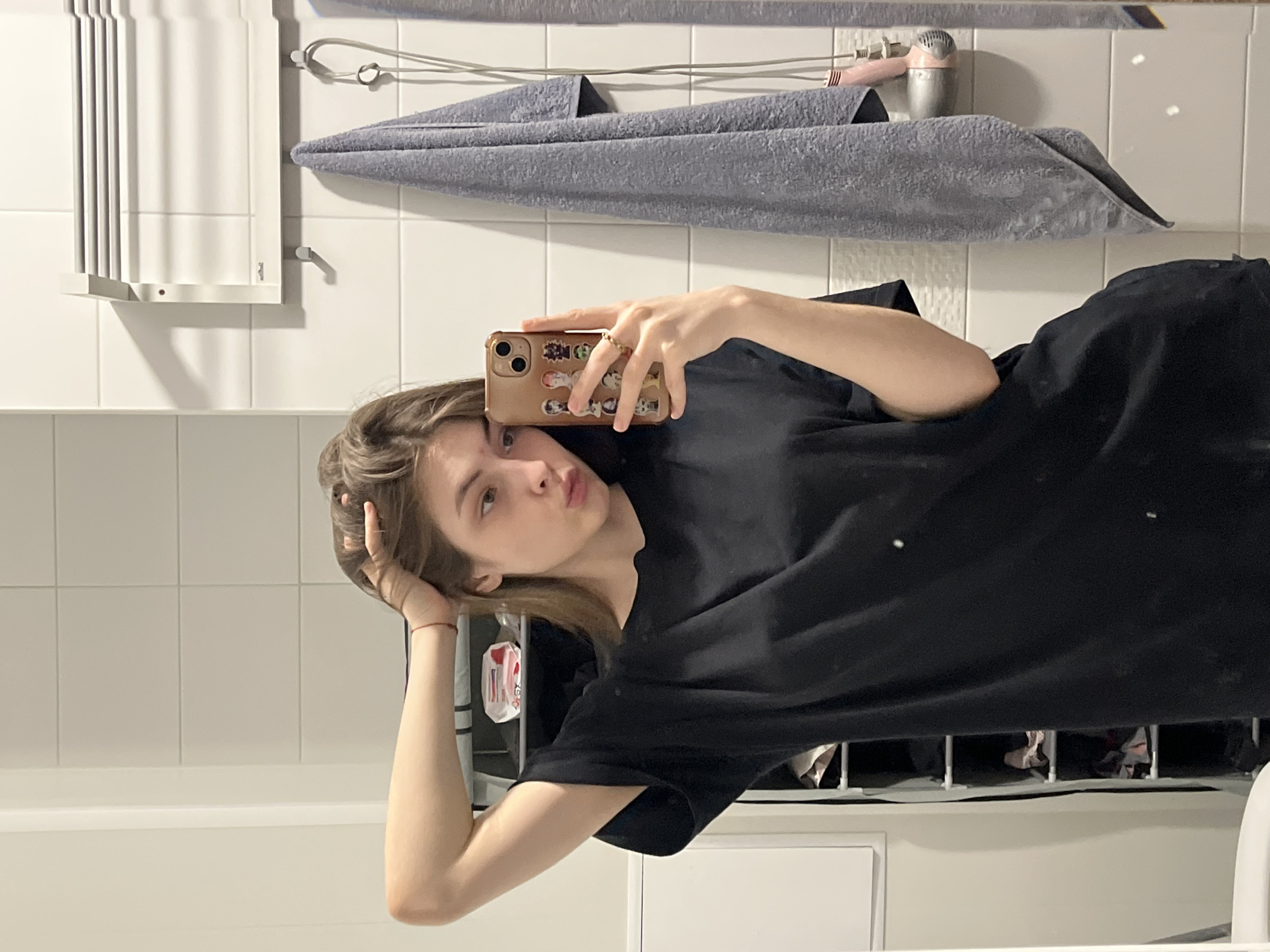 A woman is taking a selfie in a bathroom. She is wearing a black t-shirt. She is holding her phone with her right hand. Her left hand is touching her hair. There is a gray towel hanging on the wall behind her, as well as a hair dryer. The bathroom has white tiles. The location is likely indoors. There is no verifiable information to confirm that the location is in Saint Petersburg, Russia.