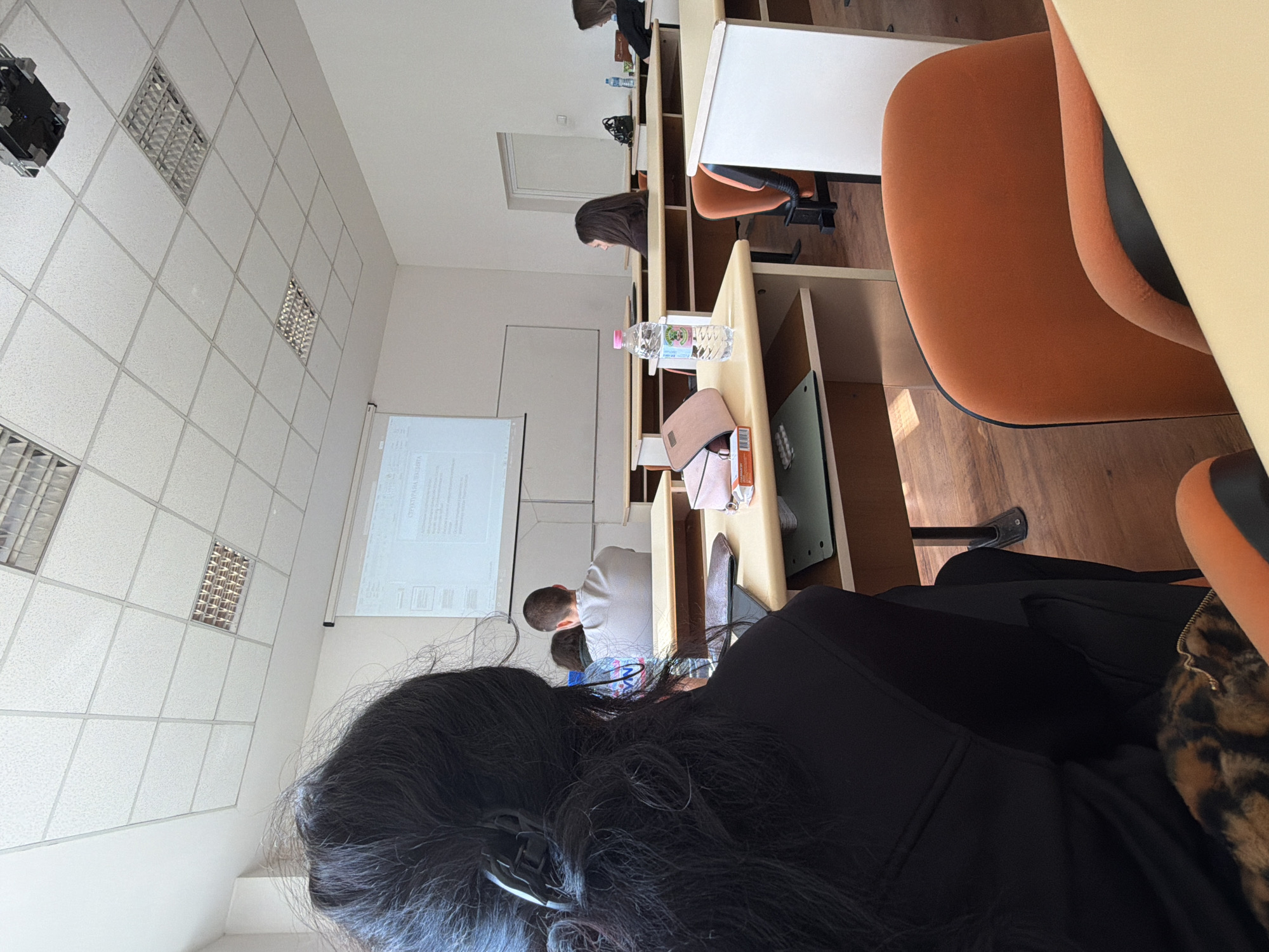 The image captures a classroom or lecture hall scene, viewed from a seated perspective looking forward. In the immediate foreground, the back of a person's head with dark, wavy hair and a black top is visible, with a glimpse of a leopard-print bag or item beside them.
Further into the room, several other students are seated at light-colored wooden desks arranged in rows. One student with short dark hair and a light-colored shirt is seen from behind, facing the front. Another student with long dark hair is visible from the side, leaning forward over their desk. In the background, another individual is faintly visible near what appears to be a window.
The students are seated in orange upholstered chairs with black bases. On one desk, a plastic water bottle and a pink/beige wallet or small bag are placed. A white projector screen at the front of the room displays a presentation. The ceiling is a white tiled false ceiling, fitted with several rectangular fluorescent light fixtures, providing ample illumination to the room, supplemented by natural light from the background window.
The activity depicted is a class, lecture, or presentation, with the students appearing attentive and focused on the content shown on the screen. The atmosphere seems academic and studious.
Visible text on the projector screen is in Cyrillic script, with the word "ОРГАНИЗАЦИЈА" (Organization) clearly discernible. The label on a water bottle on one of the desks also has text, which appears to be "GORSKA" in Cyrillic, though slightly indistinct.