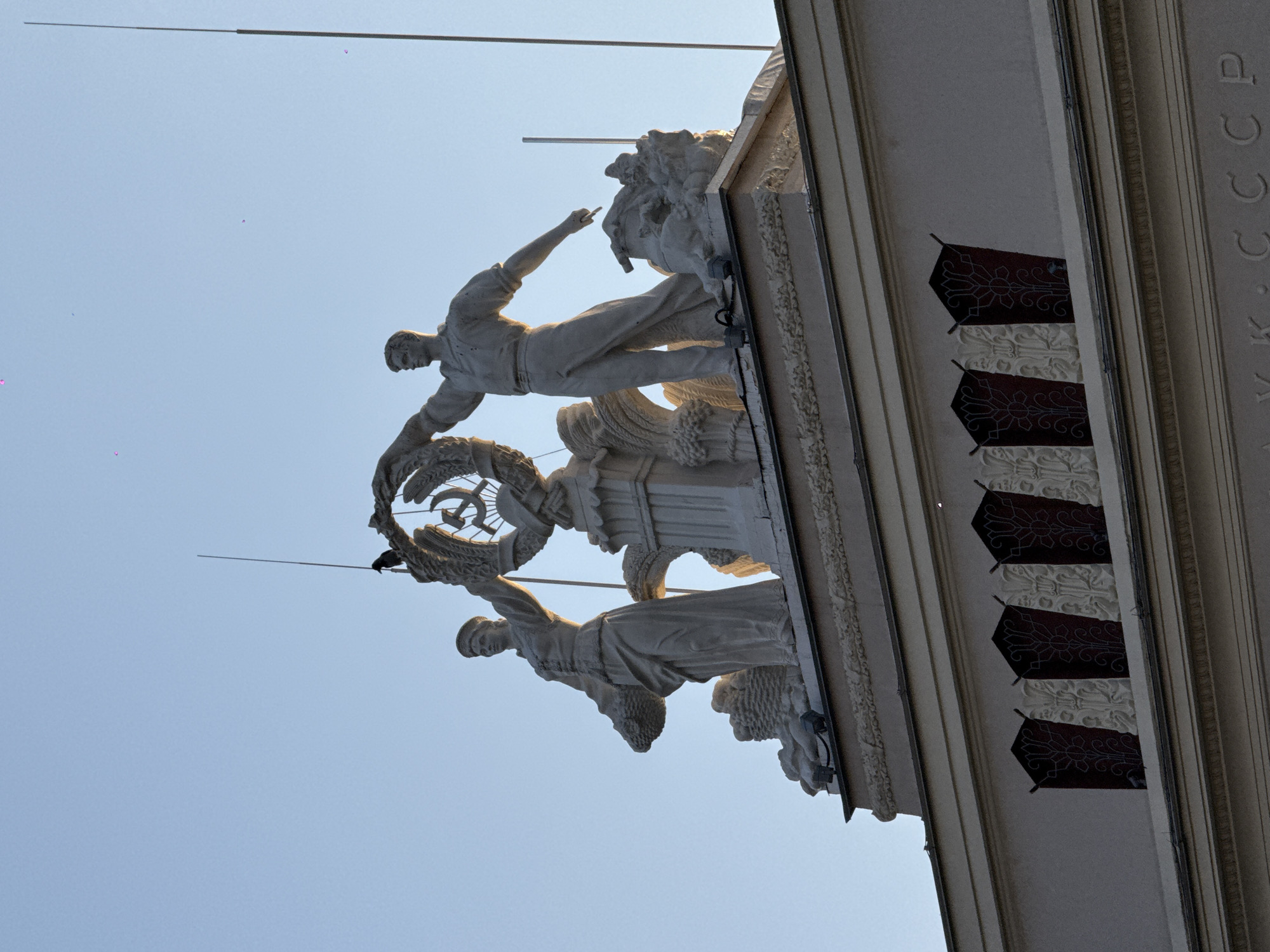 The image shows a section of the facade of the former "All-Union Agricultural Exhibition," now known as the "Exhibition of Achievements of National Economy" (VDNKh) in Moscow, Russia. The section features sculptures of a male worker and a female collective farmer holding up a wreath containing the emblem of the Soviet Union (hammer and sickle). The building is made of light-colored stone or plaster. The letters "УРСР" (Ukrainian Soviet Socialist Republic) are visible on the side of the building. The sky is clear blue.