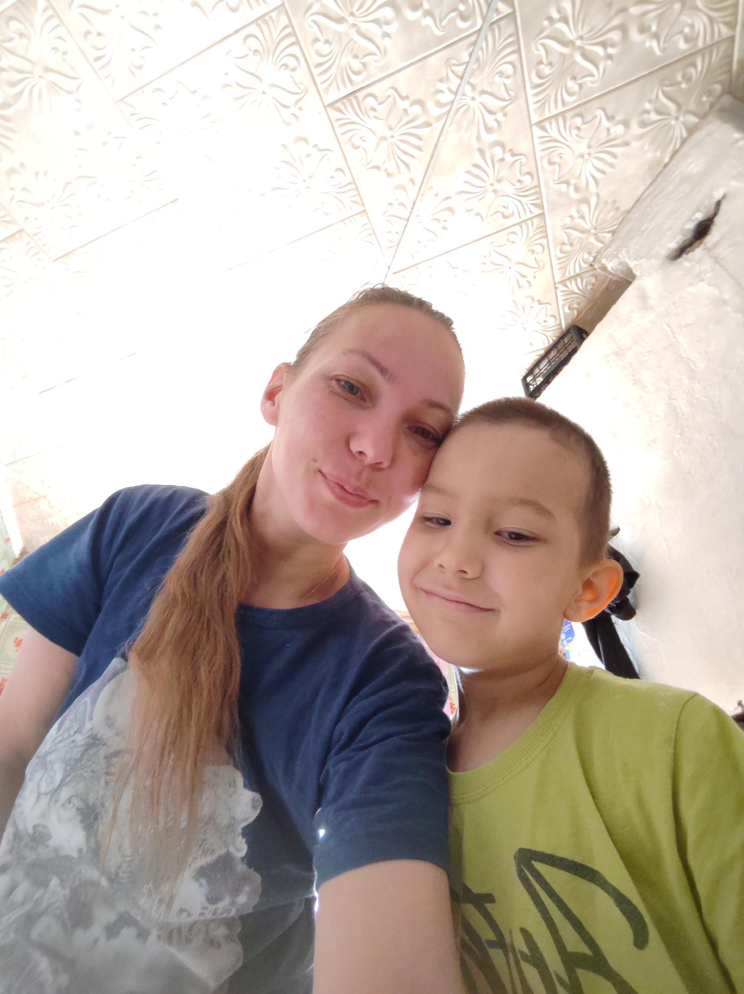 An adult female and a young male child are captured in a close-up, upward-angled selfie-style photograph within an indoor setting in the city of Чухлэм, Russia. The female, appearing to be in her 20s or 30s, has light skin, blue or green eyes, and light brown hair pulled back into a ponytail that extends over her left shoulder. She wears a dark blue t-shirt featuring a large, light-colored graphic print of an animal, possibly a wolf, on the front, and a thin gold-colored necklace. She looks directly at the camera with a subtle smile. The child, approximately 4-8 years old, has light skin and very short, dark hair. He wears a lime green t-shirt with a dark, partially visible graphic design. He is positioned to the right of the female, leaning his head against her shoulder or side, and looks slightly downward and to the right with a faint smile. The background reveals a white or off-white ceiling composed of rectangular panels, each embossed with decorative floral or scrollwork patterns, arranged in a diagonal pattern. To the right, a textured, light-colored wall is visible, featuring a dark, rectangular object with horizontal slats mounted on it, and indistinct dark items below. The scene is brightly illuminated, casting significant light on the ceiling.