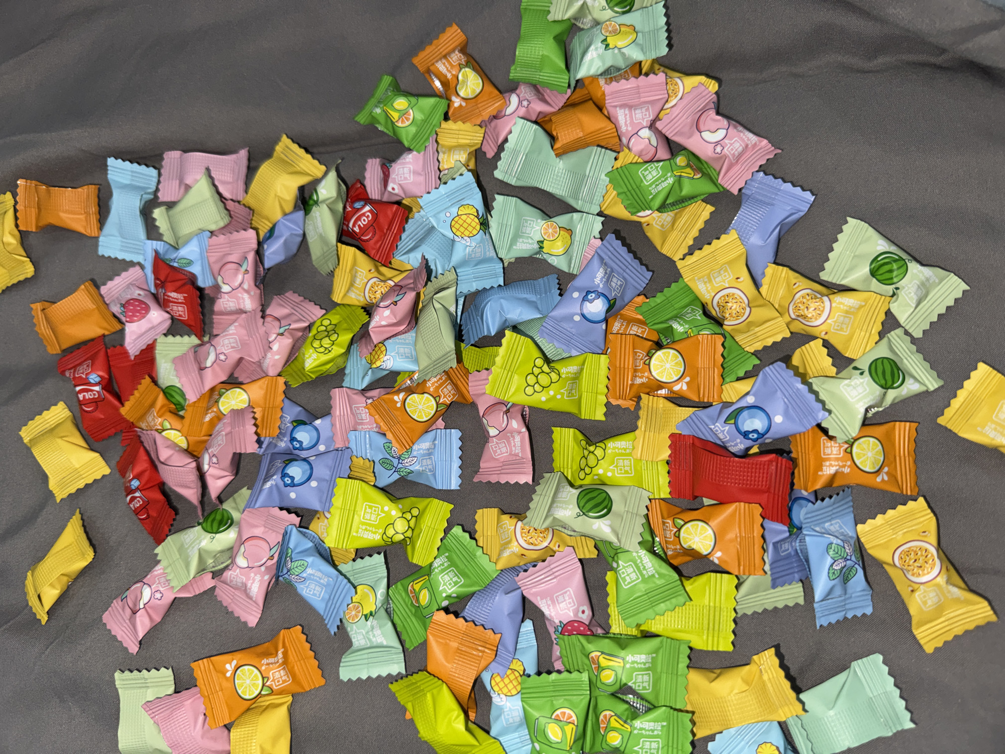 The image displays a collection of individually wrapped candies scattered on a gray surface, likely fabric. The candies come in various colors and feature fruit-themed designs, including pineapple, watermelon, orange, and lemon. Some wrappers also have the word "COLA" printed on them. The candies are seemingly randomly distributed, creating a colorful array on the gray background. The wrappers have the phrase "小可奥拉 かーちゃんおら" in Japanese. The location is unknown and cannot be determined from the image.
