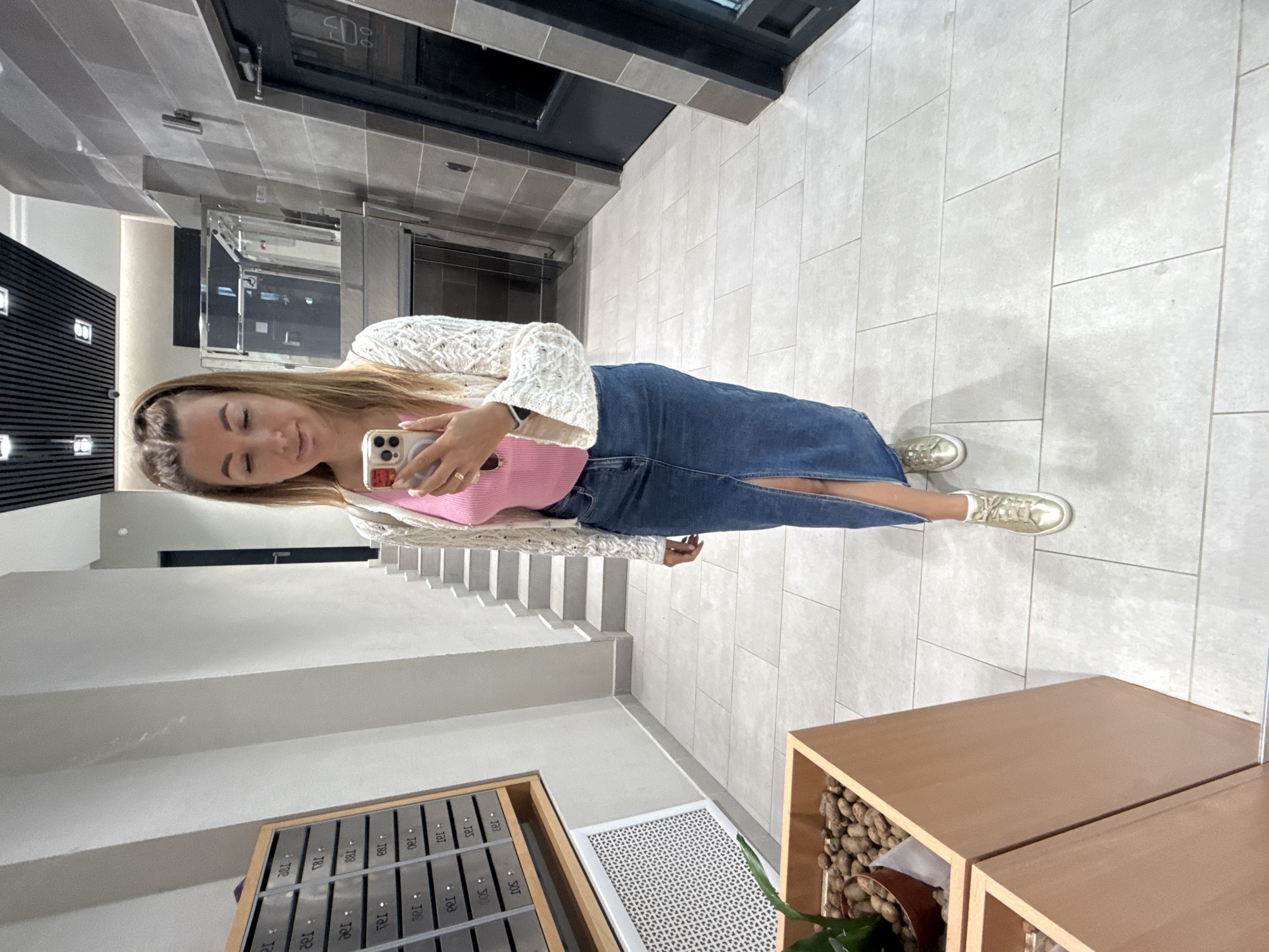 A woman with blonde hair, wearing a pink top, a white knitted cardigan, and a long denim skirt, takes a mirror selfie in a hallway. She is holding a smartphone in her right hand and wears a ring on her left ring finger. Her left foot is slightly in front of her right. To her left, there are mailboxes mounted on the wall and a wooden planter with a plant and pebbles. Ahead of her, a staircase ascends to the left, and to the right, there are doors and a tiled wall. The floor is tiled with large, light-colored square tiles. The lighting appears to be artificial.