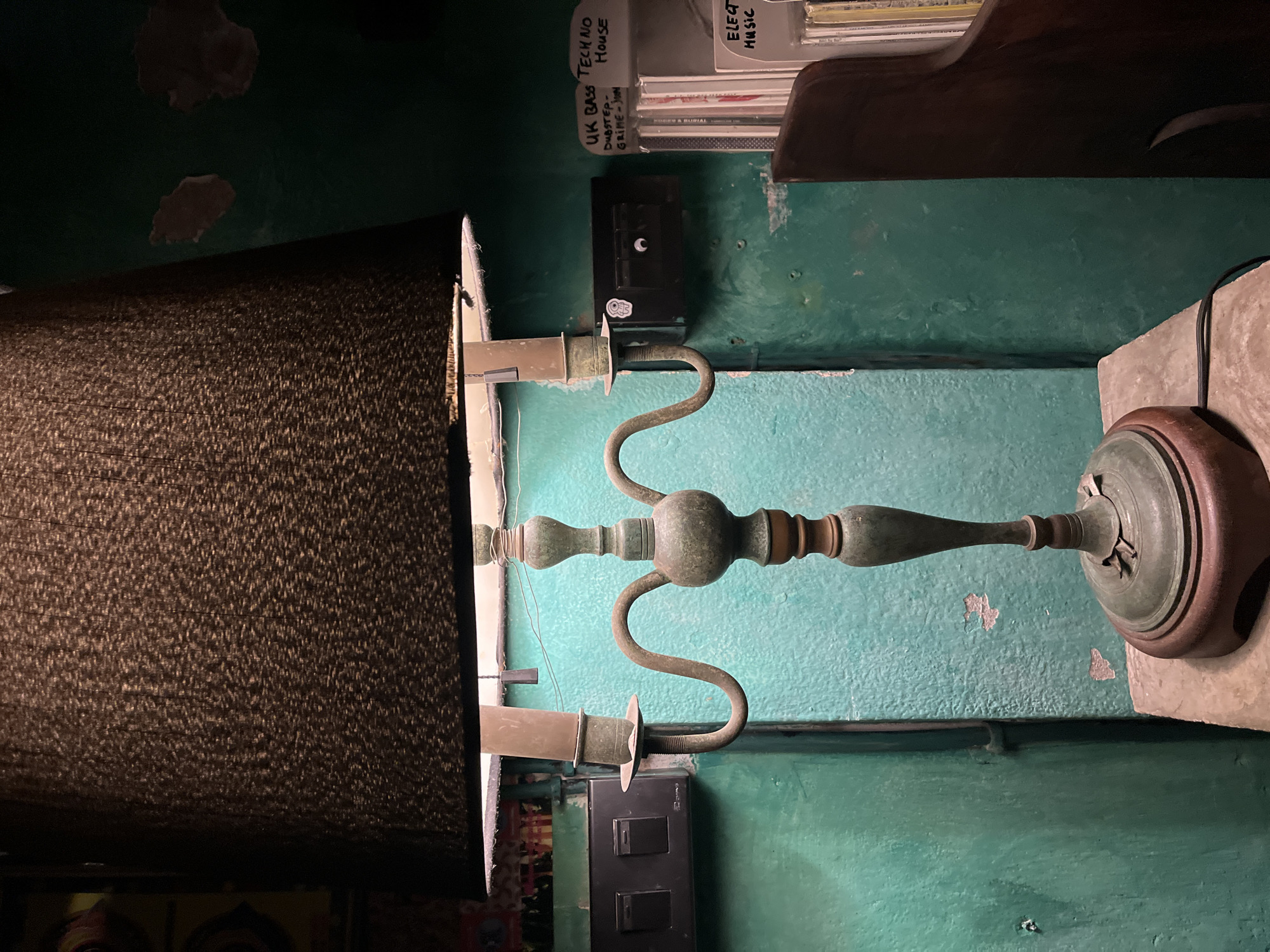 A photograph taken indoors in Chiang Mai City Municipality, Thailand shows a floor lamp with a dark brown lampshade dominating the left side of the frame. The lamp has a weathered, green-patina metal stand with curved arms holding the light sockets. The wall behind the lamp is painted turquoise with visible wear and paint chipping. Above the lamp, a dark shelf holds a collection of music CDs, labeled with genres like "UK Bass," "Techno House," and "Electronic Music." Two black electrical outlets are mounted on the wall, one on either side of the lamp stand. The lighting is dim, and the overall atmosphere suggests a lived-in, possibly vintage, space.