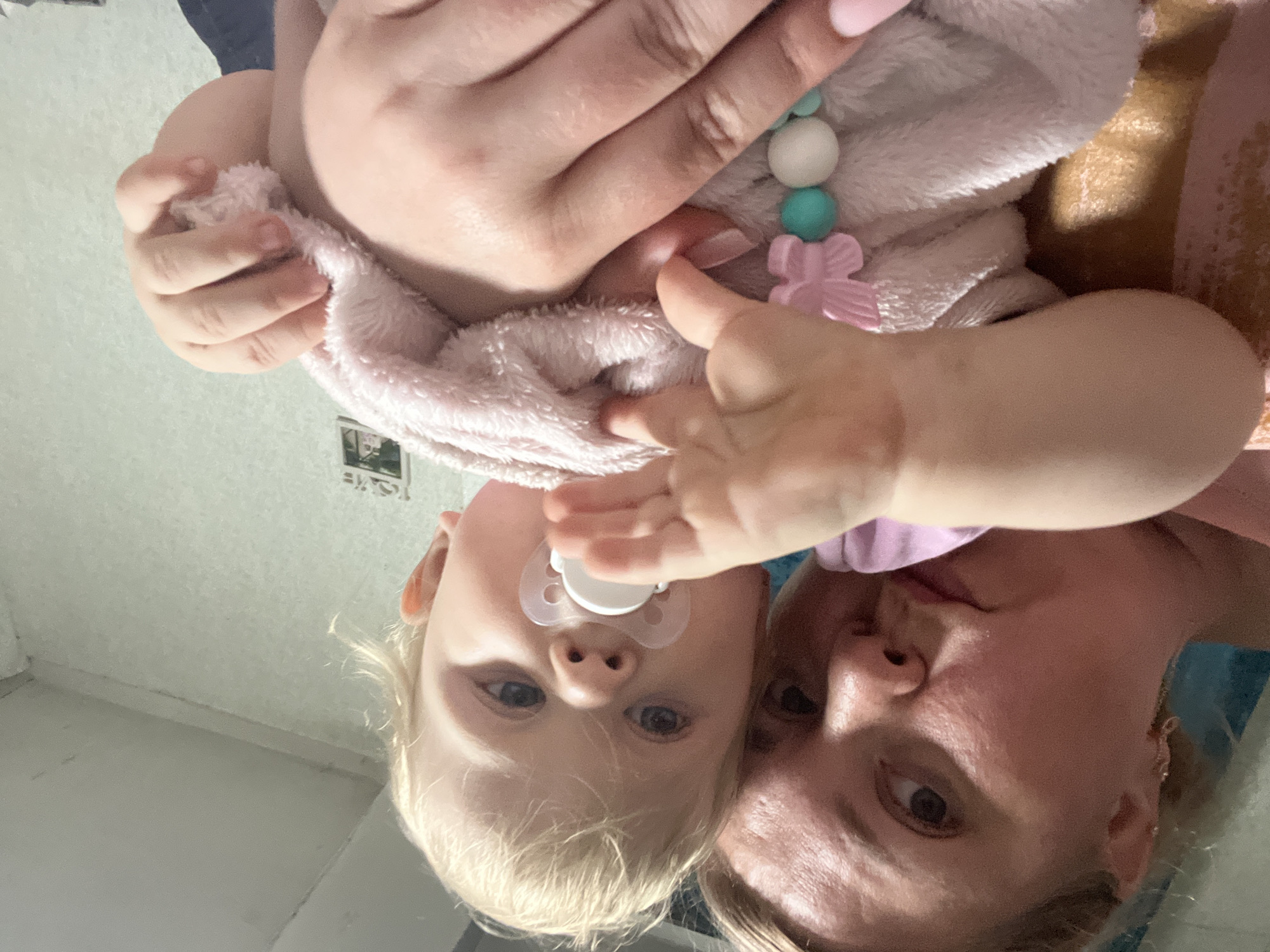 A woman and a baby are being photographed. The woman is holding the baby, who has a pacifier in their mouth. The baby is wearing a pink blanket. The background appears to be a room. The image was taken in the city of Фоки, Russia.