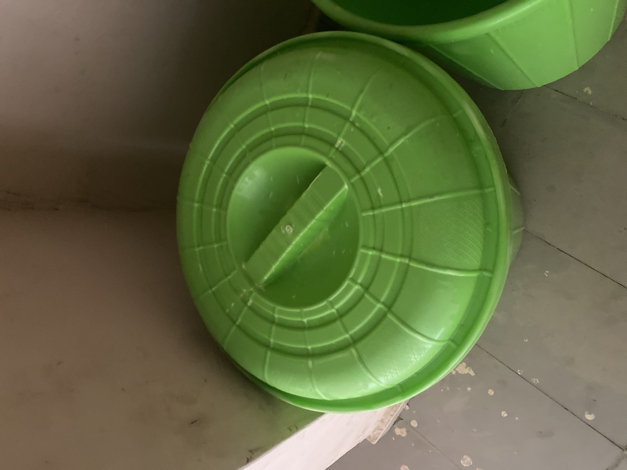 In Bulamare, Nigeria, a bright green plastic bucket with a matching lid sits on a weathered, grey floor with scattered white debris. The lid features concentric ridges and a raised handle in the center. Another green bucket is partially visible to the right of the main subject. The background consists of a light brown wall meeting a white partition. The lighting is diffused, casting soft shadows.
