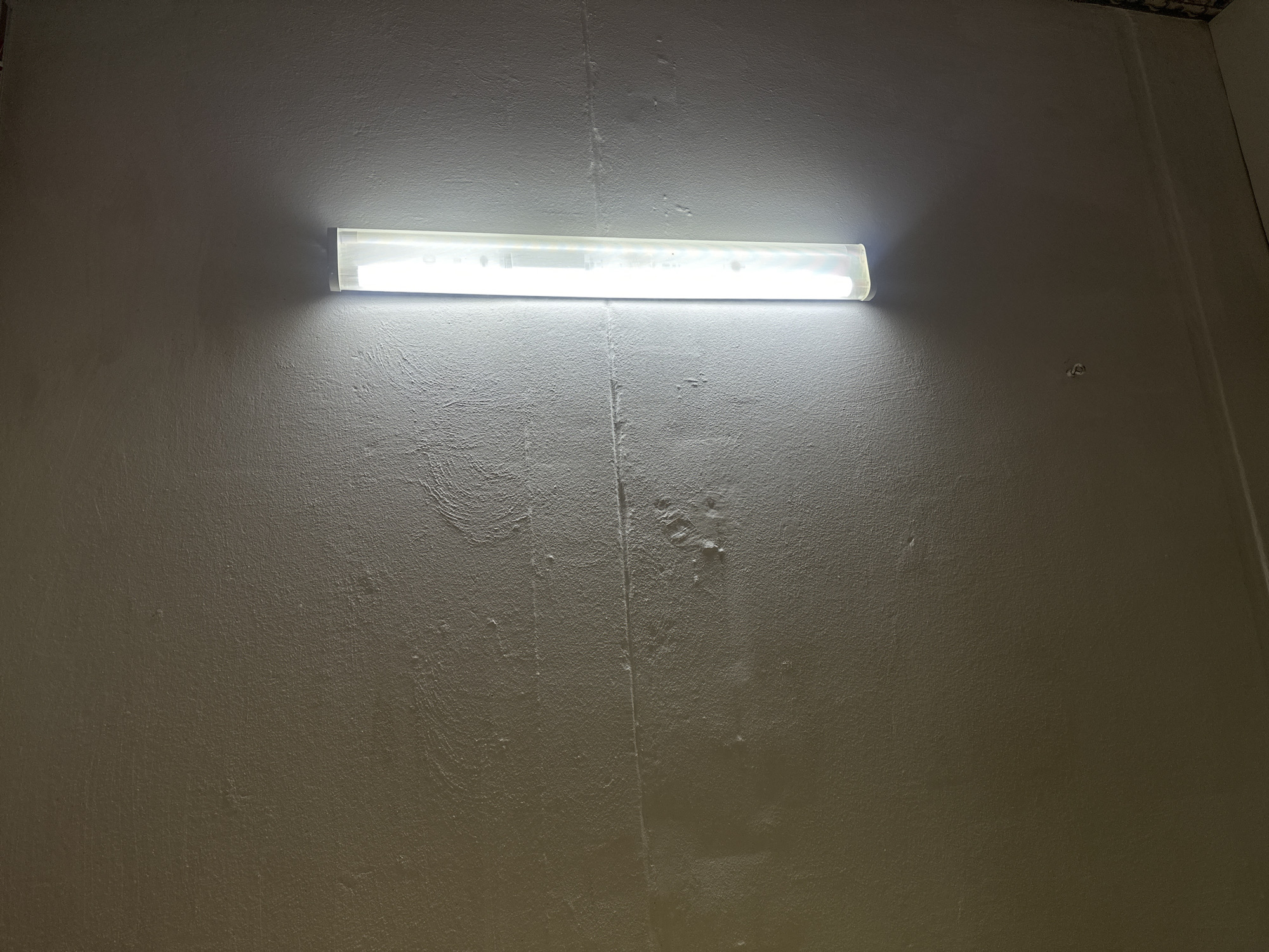The scene, located in Aktanysh, Russia, displays a single, vertically oriented tubular light fixture affixed to a textured, light grey wall. The fixture emits a bright white light, intensely illuminating the immediate wall surface and creating a luminous halo that gradually fades into darker areas of the wall. The wall exhibits an irregular, rough plaster finish. A distinct horizontal seam or crack is visible across the wall approximately at its mid-height. Below this seam, several irregular bulges and minor depressions are present on the wall's surface. No individuals, animals, or specific actions are depicted. The environment is an unadorned interior space.
