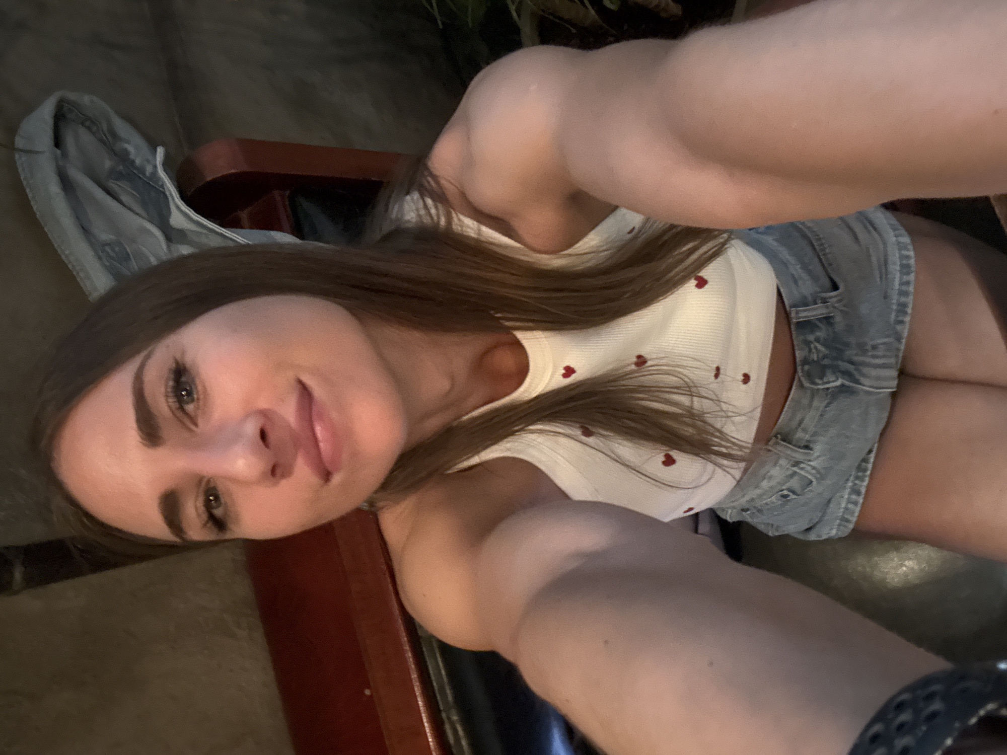 The media file shows a young woman taking a selfie. She is wearing a white top with small red hearts and denim shorts. Her long brown hair is falling over her shoulders. She is sitting on a dark wooden chair with a piece of folded denim clothing draped over its back. The background is dimly lit, with hints of greenery, suggesting an indoor setting possibly at dusk or in the evening. The woman's expression is relaxed and she appears to be smiling faintly. There are no visible text elements in the image. The overall impression is casual and personal.