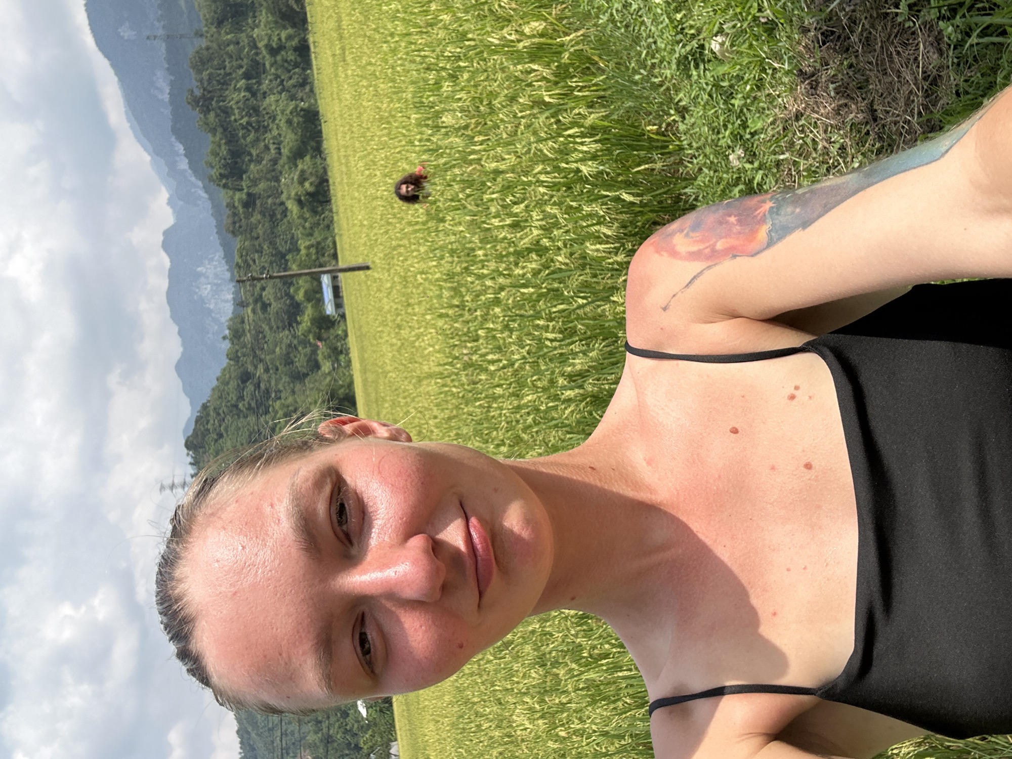 A selfie, taken in portrait orientation but displayed rotated, captures a young adult female in a vast natural setting under a partly cloudy sky. The main subject, positioned in the foreground, looks directly at the camera with a faint smile. Her fair skin appears slightly reddened from sun exposure, and her light hair is pulled back. She wears a black strap top, revealing her left shoulder and arm, which bears a vibrant, abstract tattoo in shades of orange, blue, and black. Several moles are visible on her chest and shoulder.
Behind her, an expansive field of tall, vibrant green crops, likely rice, stretches into the middle ground. Partially obscured by the crops, the head and shoulders of a second person are visible in the middle distance. A wooden pole with a small, unreadable blue sign is also present in the field.
The background features multiple layers of majestic mountains and hills, densely covered in green vegetation. The most distant mountains appear hazy and blue against a sky with scattered white and gray clouds. The bright sunlight suggests it is late morning or afternoon. The scene conveys a peaceful, rural atmosphere, consistent with locations like Ngọc Đường Commune, Vietnam. No readable text is visible on the small blue sign.