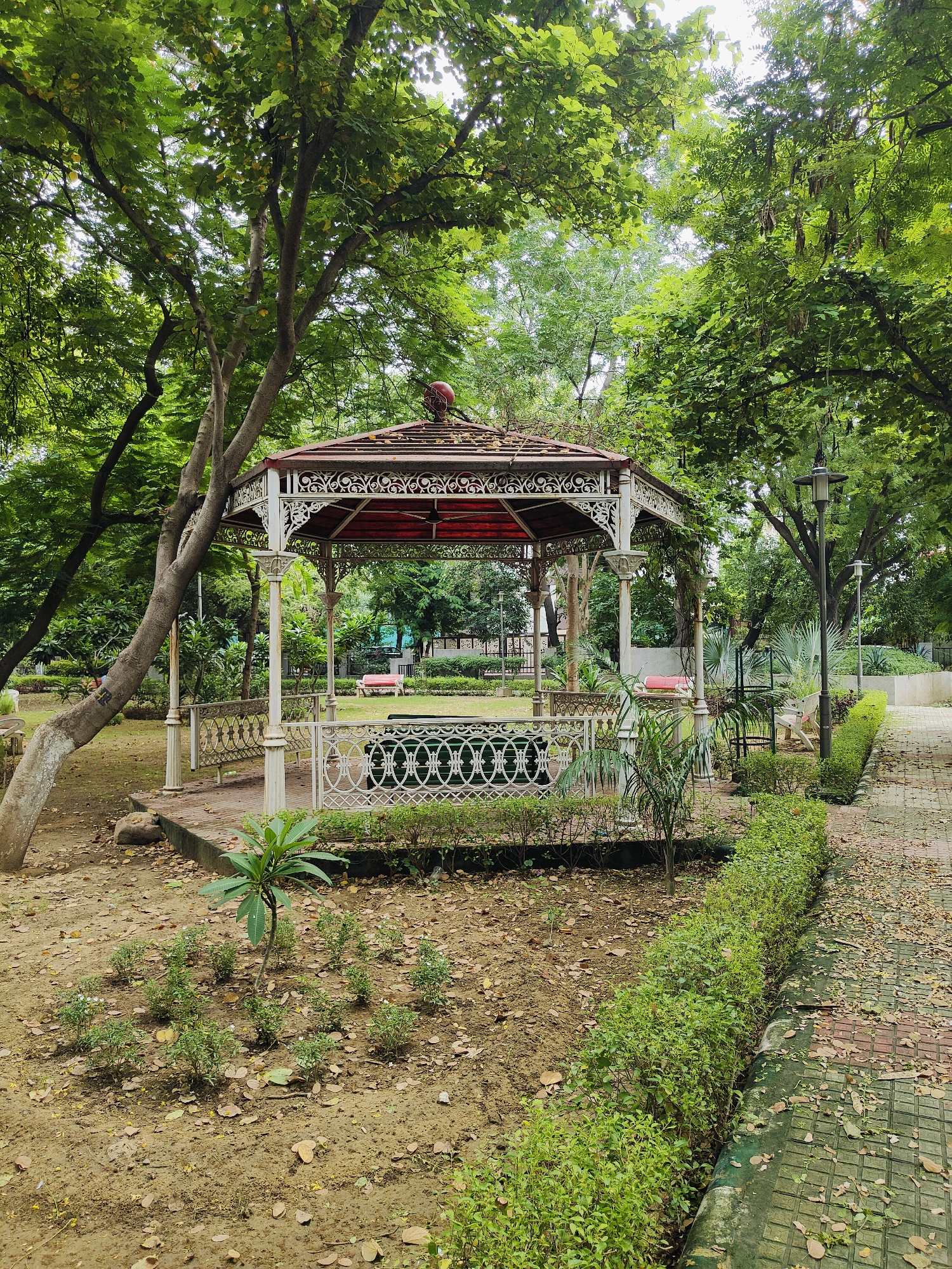 The image depicts a gazebo in a park, likely located in Gurgaon, India. The gazebo has a white ornate metal structure with a red roof and is surrounded by lush green trees and foliage. A paved pathway with a brick pattern is visible to the right of the gazebo, bordered by a manicured hedge. The ground is covered with fallen leaves, suggesting an outdoor, natural setting. There are no people visible in the scene, and the environment appears to be peaceful and well-maintained. The overall impression is of a serene public space within a city.