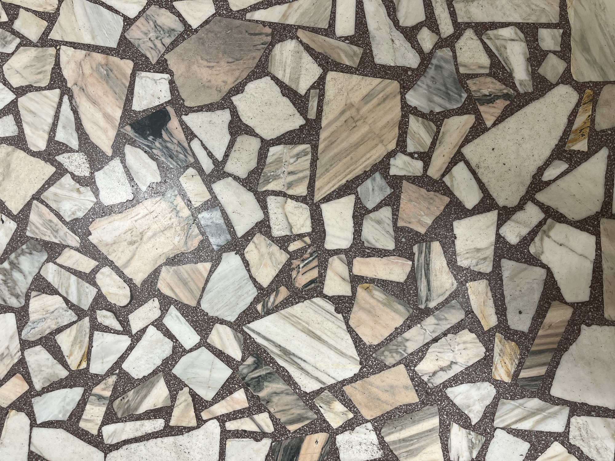 The image provides an overhead, close-up view of a floor made from a composite material, resembling terrazzo or a mosaic of natural stone. The surface is characterized by numerous irregularly shaped and sized stone fragments, predominantly marble or a similar material, embedded within a darker, granular matrix.
The stone pieces display a diverse palette of light colors, including white, off-white, cream, and various shades of light gray. Many fragments feature distinct veining patterns in contrasting tones such as darker gray, black, light brown, and subtle pinkish or yellowish hues, contributing to the material's natural aesthetic. A few isolated pieces exhibit more prominent dark gray or black marbling.
The binding material, acting as the grout, is a dark brown to reddish-brown color and appears finely speckled with tiny lighter particles. This darker matrix forms an irregular network that outlines each stone piece, creating a stark contrast with the lighter-colored fragments.
The overall pattern is organic and non-repetitive, resulting from the random arrangement and varied forms of the stone components. The surface appears smooth, likely polished, and is illuminated by an even, diffused light, suggesting an indoor setting. There are no visible signs of significant wear, damage, or discernible text. The image solely captures the textural and chromatic details of this decorative flooring.
