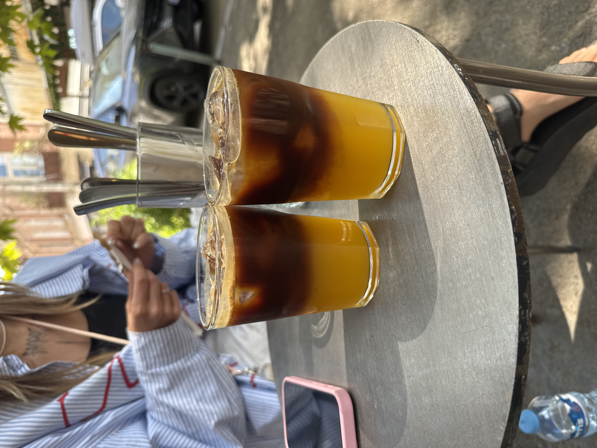 Two glasses filled with an orange beverage and dark liquid topped with ice are placed on a round metal table. The glasses are positioned in the foreground, with a pink-cased smartphone to the left. In the background, a person wearing a blue and white striped shirt is visible, holding what appears to be a receipt. A metal cup containing silverware is behind the glasses. The scene appears to be outdoors, with cars and buildings visible in the background. The lighting suggests it is daytime, possibly in the city of Tbilisi, Georgia, given the architectural style of the buildings and the general ambiance.