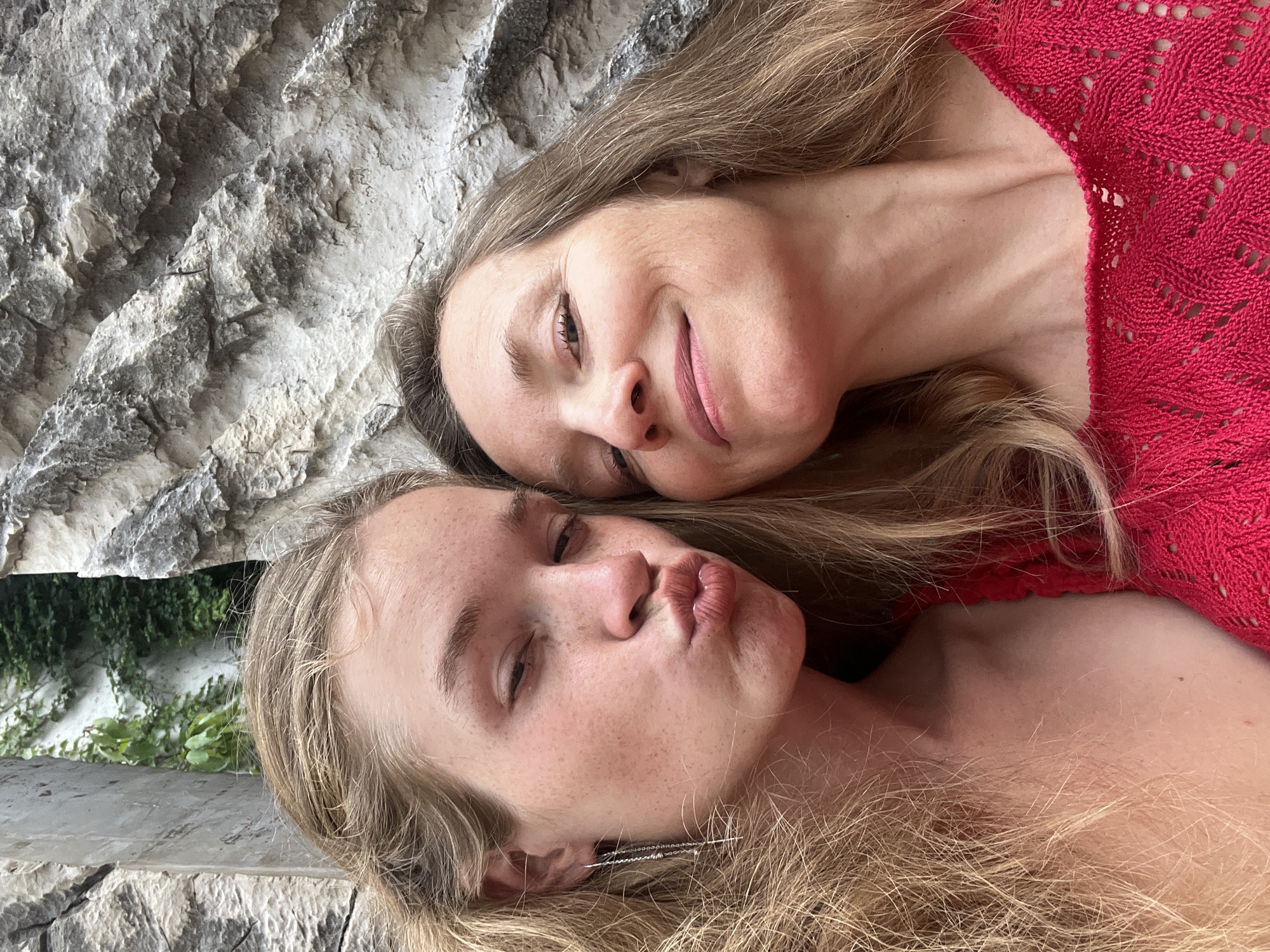 This is a selfie featuring two individuals, likely a mother and daughter, in Budva, Montenegro. The person on the left, with long blonde hair and freckles, is puckering their lips. The person on the right, with brown hair, is smiling. They are positioned in front of a stone wall with greenery visible in the background. The overall impression is one of a casual, outdoor portrait.