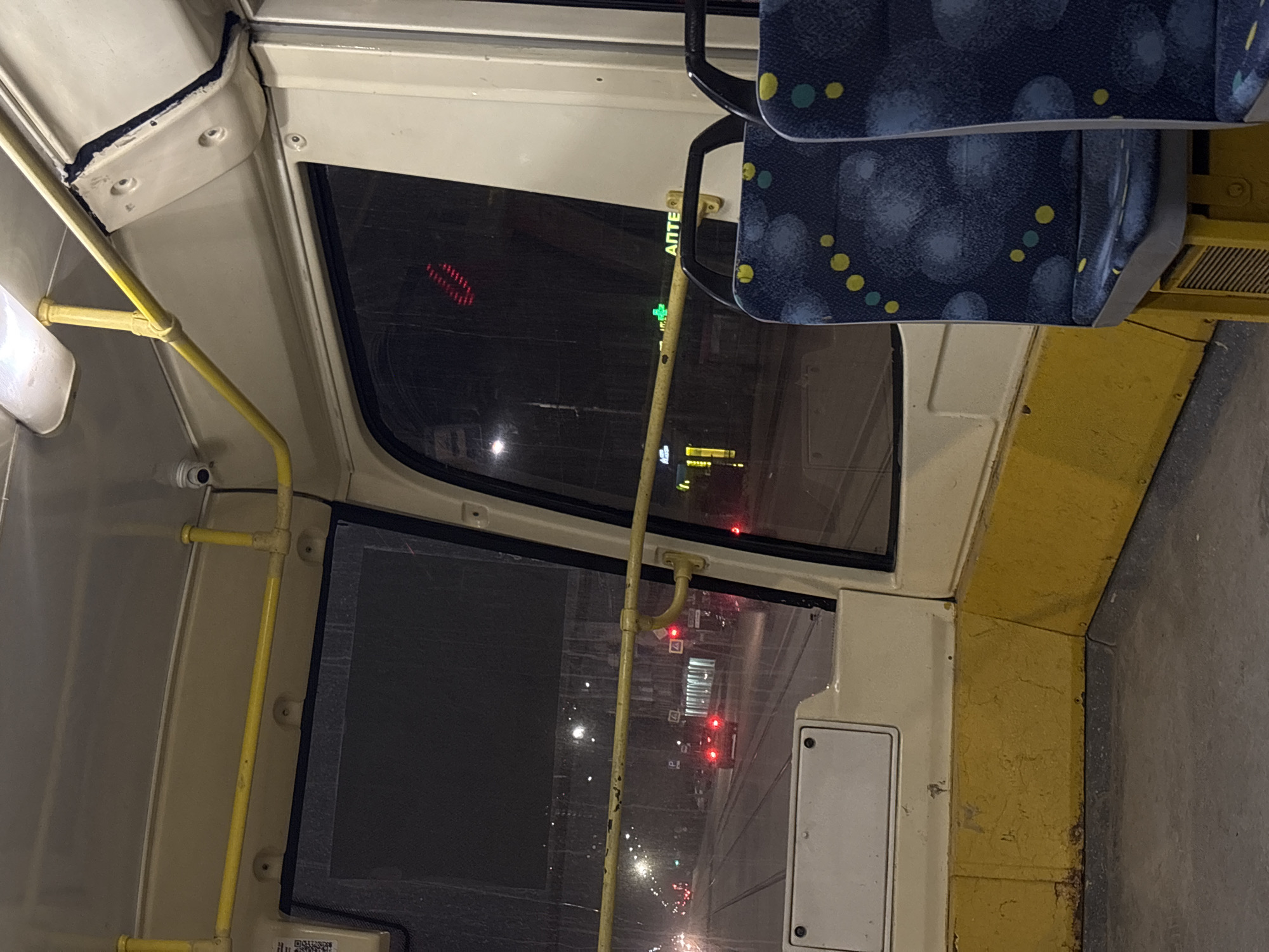 The image depicts the interior of a bus, likely in Perm, Russia, as suggested by the АПТЕКА sign visible through the window. The interior features a yellow handrail, window, and a blue patterned seat. Outside the window, there are street lights, traffic lights, and buildings visible, indicating it is nighttime. There are no people visible in the image. The focus is on the bus's interior and the view through the window.