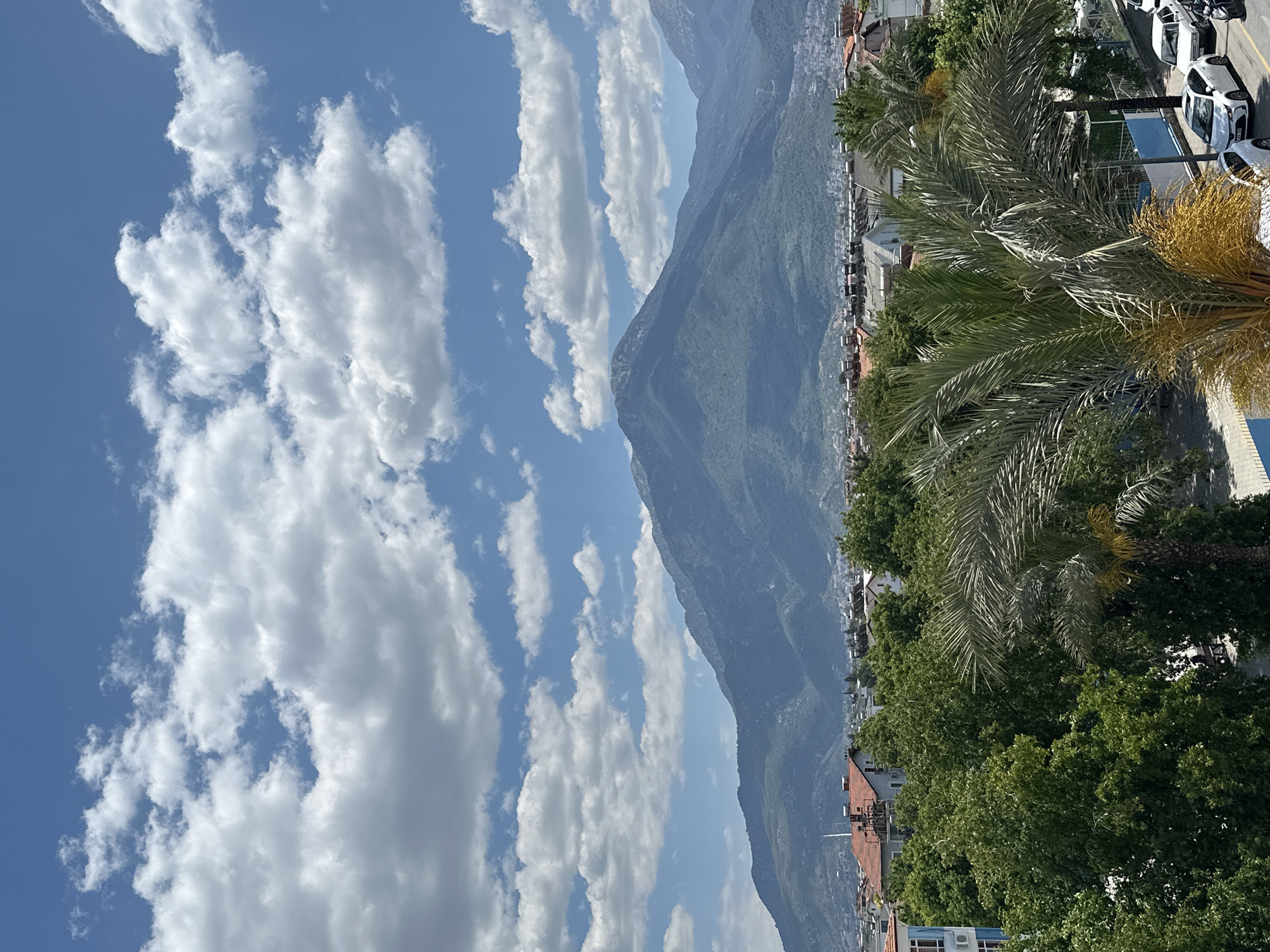 A vertical, eye-level photograph depicts a daytime scene in Fethiye, Turkey. The sky, predominantly blue with scattered white cumulus clouds, occupies the left half of the frame. A large, shadowed mountain range runs down the center of the photograph.
On the right side of the mountain range and running down to the right edge of the frame, a portion of the city is visible, including residential buildings with red tile roofs, interspersed with green trees. The top right corner of the image shows a parking area with several white cars and some yellow objects. A palm tree with green and yellow fronds dominates the foreground on the right, partially obscuring the cityscape.