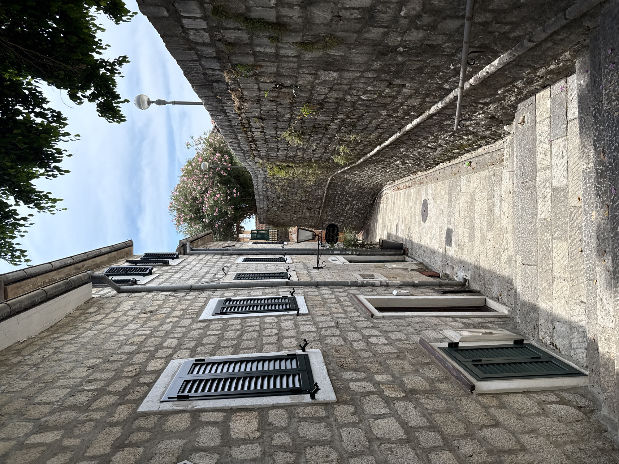 This media file depicts a charming, narrow street in Herceg Novi, Montenegro, likely during the daytime under clear skies. The scene is dominated by traditional stone architecture. A building with rough-hewn stone walls and multiple dark-shuttered windows lines the left side of the street, viewed from a low angle looking upwards. The windows appear to be uniformly designed, adding to the architectural consistency.
The street itself is paved with light-colored stone, and it curves upwards and away from the viewer, suggesting a hilly terrain common in coastal Montenegrin towns. To the right, the street transitions into a set of stone steps, also paved with large, flat stones. A stone wall, constructed from smaller, more uniform blocks, forms the upper boundary of the street on the right side, with greenery and moss growing in its crevices.
A lamppost with a modern, rounded fixture stands prominently against the bright blue sky, partially obscured by the dense green foliage of a large tree on the left. Further down the street, a tree with delicate pink blossoms adds a touch of color to the scene. The overall impression is one of old-world charm and tranquility, typical of historic Mediterranean towns. No people are visible, and the focus is on the architectural elements and the natural surroundings. The lighting suggests it is midday or early afternoon, with bright sunlight casting some shadows.