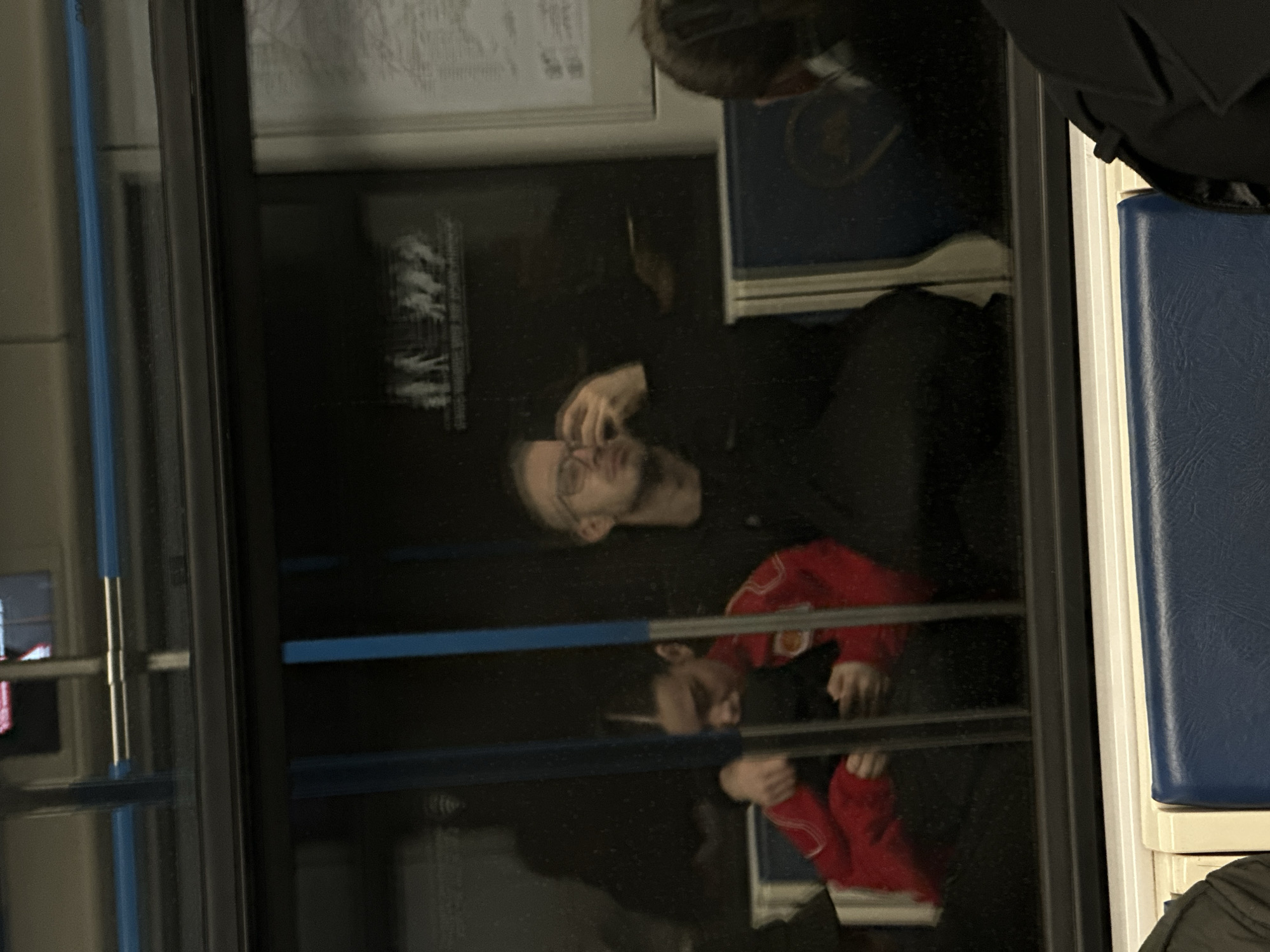 The image depicts the reflection of a man with glasses on a train in Moscow, Russia. The man is wearing a black shirt and a red jacket with white cuffs. He is looking at his reflection and adjusting his glasses. The train's interior includes blue cushioned seats. A public transport information poster is visible above. The scene appears to be inside a metro carriage.
