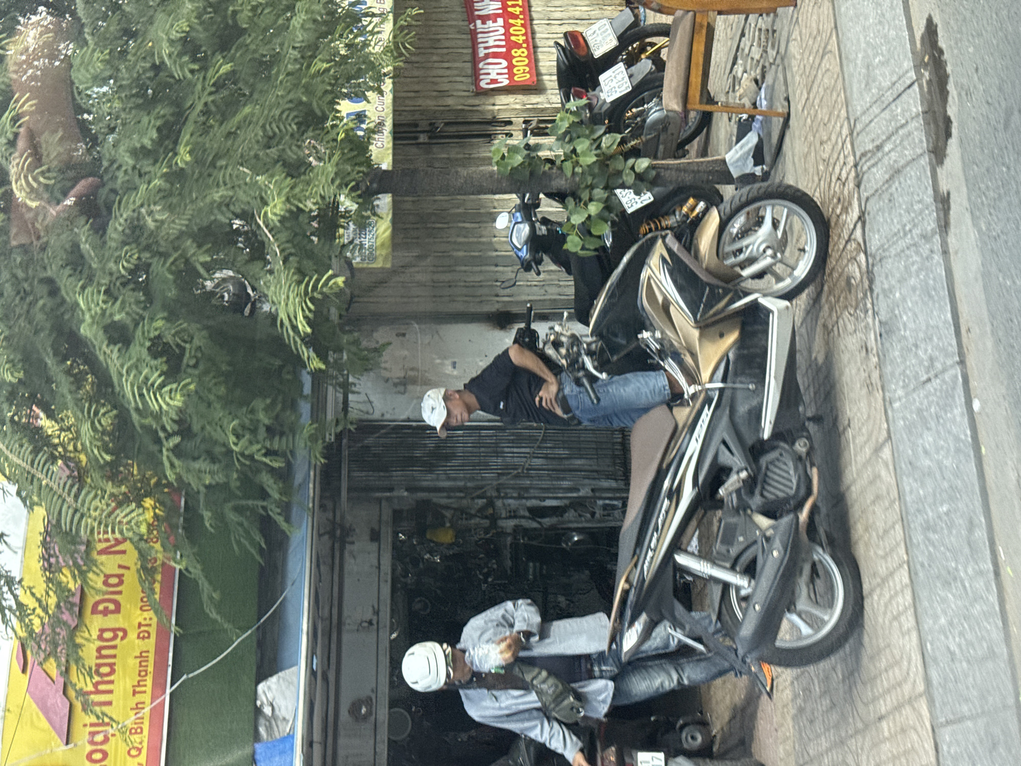 The image depicts a street scene in Thủ Đức, Vietnam, likely in front of a shop or workshop. In the foreground, a man wearing a white helmet, sunglasses, and a light-colored jacket stands next to a motorcycle. He is holding a clear plastic bag with some contents inside. To his right, another man in a white cap and dark t-shirt stands with his hands on his hips, looking towards the left. Several other motorcycles are parked behind them. A large tree with green foliage extends into the upper part of the frame, partially obscuring a yellow sign with Vietnamese text. A red sign with white text, also in Vietnamese, is visible on the right side of the image, with the number "0908.404.41" displayed prominently. The ground is paved with patterned tiles, and a gray concrete curb runs along the edge of the road. The overall impression is a busy urban environment, possibly a commercial area where motorbikes are a common mode of transportation and business.