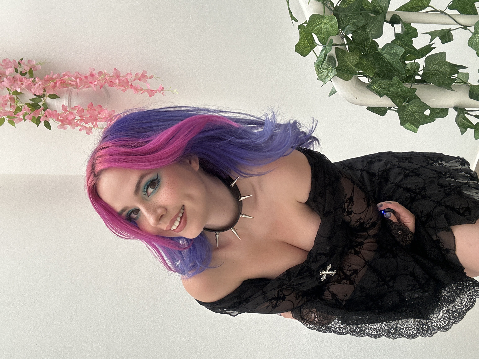 A young woman with vibrant, two-toned hair, pink blending into purple, is captured in a close-up, sideways-oriented portrait. She smiles broadly at the viewer, revealing white teeth. Her face features light blue eyeshadow, defined eyelashes, and scattered freckles across her nose and cheeks. She is wearing a black, sheer, off-the-shoulder dress with intricate lace patterns and a scalloped lace trim along the hem. A prominent black choker with sharp silver spikes adorns her neck, and a small silver cross charm is pinned to her dress near her chest. In her left hand, a small, blue, possibly glowing object is visible.
The setting appears to be indoors, against a plain white wall. The background is minimally decorated with artificial plants. On the left side, a cluster of delicate pink cherry blossom-like flowers is arranged, seemingly hanging or affixed to the wall. On the upper right, green artificial ivy vines are wrapped around a white pipe or beam, adding a touch of natural accent to the stark white space. The overall impression is one of a cheerful and stylish pose.