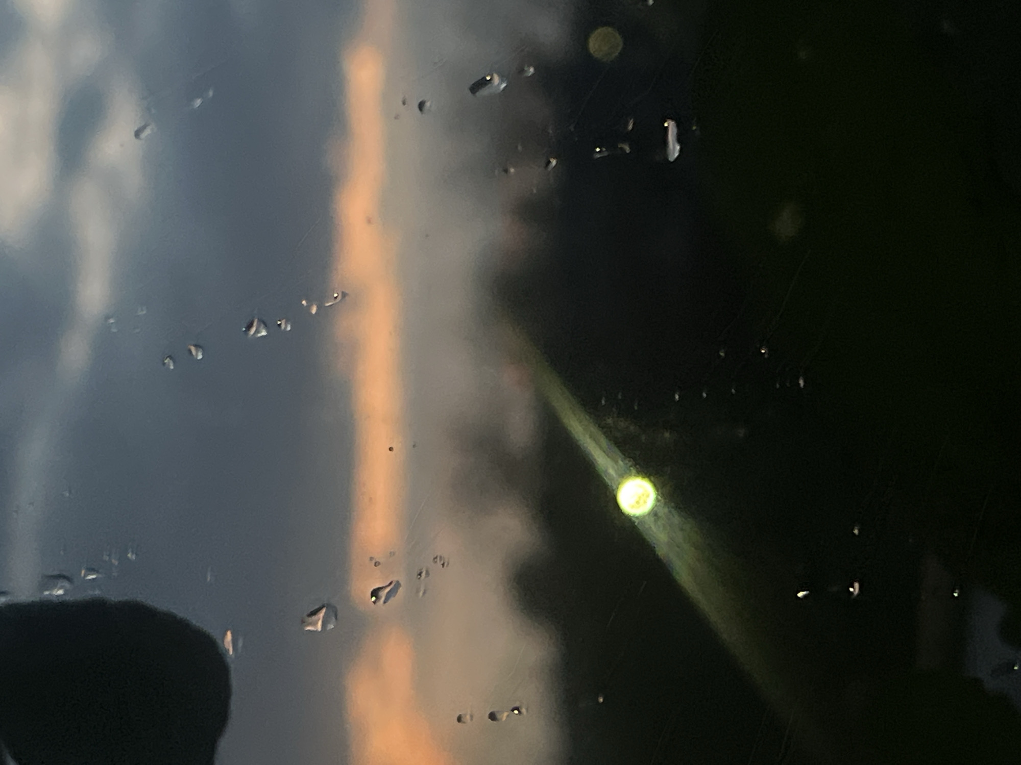 The image shows a view through a wet window. Raindrops are visible on the glass surface. Outside, there is a dark area, possibly indicating night or heavy shadows. To the left, the sky is visible, showing a mix of grey and orange hues, suggesting either sunset or sunrise. A bright, blurred light source is present, potentially a streetlight or another artificial light. The specific location within Votkinsk, Russia, is not discernible from the image. No people are visible.