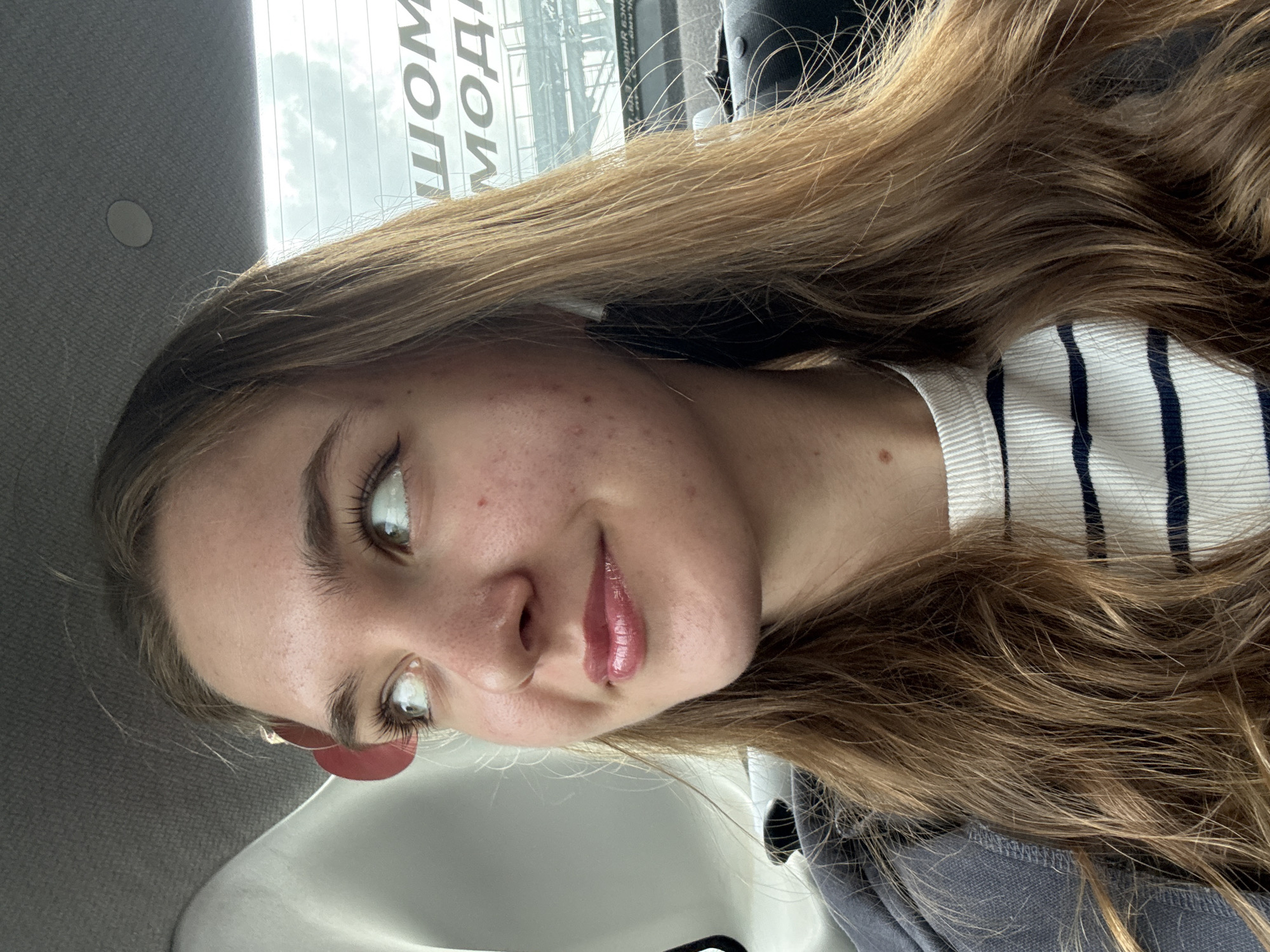 A young woman with light brown hair is the central subject of the image. She has fair skin, light eyes, and is wearing a striped top. She's in what appears to be a vehicle, given the car interior elements visible (headliner, window frame). In the background, through the window, is a partially obscured urban scene, possibly indicating the city of Perm, Russia, as text is visible on the window in the Russian language. The overall composition suggests the woman is taking a selfie or being photographed from a close distance within the vehicle.