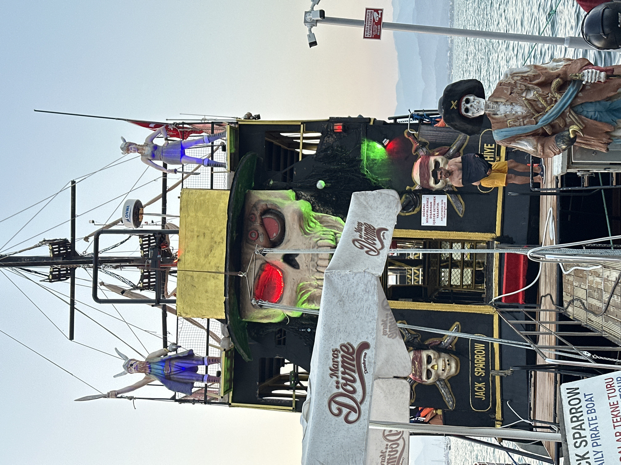 This is a medium shot of the side of a pirate-themed boat, likely a tour boat, named "Jack Sparrow." The boat is decorated with skeletal figures and pirate imagery. In the foreground, two large, stylized skeletons are visible, one wearing a pirate hat and the other part of the boat's facade. A large, illuminated skull with glowing red eyes and green neon accents is prominently displayed. There are also smaller figures dressed as pirates or warriors, some with blue lighting.
The boat is moored at sea, with a body of water and distant hills or mountains visible in the background, suggesting a coastal location. The sky is clear and bright, indicating it is daytime.
Several pieces of text are visible. A sign reads "JACK SPARROW DAILY PIRATE BOAT" and "FETHİYE TURU." Another sign in Turkish states that outside drinks are prohibited and thanks visitors for their understanding. There is also a sign warning that the area is being recorded by cameras. Additionally, the word "Maras Dövme Dondurma" appears on paper wrapping, likely from an ice cream vendor.
The overall impression is a festive, possibly tourist-oriented attraction designed to evoke a pirate adventure. The decorations suggest a playful and slightly spooky theme.
