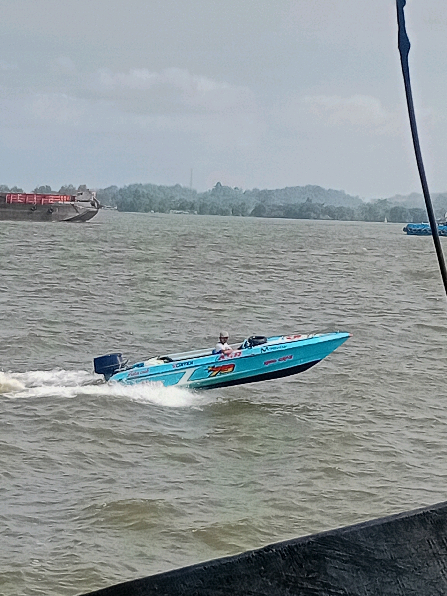 A high-speed blue and white motorboat with a single occupant is speeding across a body of water. The boat has graphics and text on its hull, including the word "CONEX" visible on the side. The occupant, wearing a white shirt and a light-colored cap, appears to be operating the boat. The boat is creating a significant wake, indicating its speed. In the background, a large barge with red cargo is visible on the water, and a tree-lined shore with a hazy sky is discernible. The overall scene suggests a recreational or competitive activity taking place on the water near a populated area. This location is identified as Sungai Meriam, Indonesia.