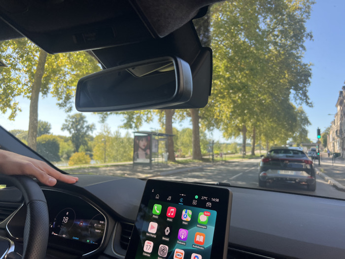 A forensic view from the interior of a modern vehicle, likely a Renault, looking forward through the windshield on a clear, sunny afternoon in Orléans, France. The digital instrument cluster displays "18" and "Comfort" mode. A large infotainment touchscreen on the right shows a grid of applications, "27°C", and the time "14:58". A light-skinned hand rests on the dashboard's left side. The rearview mirror is centered at the top.
Outside the vehicle, a two-lane road stretches ahead. A dark-colored SUV is observed directly in front, stationary or moving slowly. To the right, traditional European multi-story buildings line the street, and a green traffic light is visible in the distance. To the left, a row of mature deciduous trees with lush green foliage borders the road, beyond which a wide body of water, identifiable as the Loire River, flows. On the embankment between the road and the river, a modern bus stop shelter features an advertisement poster of a young woman. The sky is clear blue. The scene is consistent with driving along one of the quays bordering the Loire River in Orléans, such as Quai du Châtelet or Quai Cypierre.