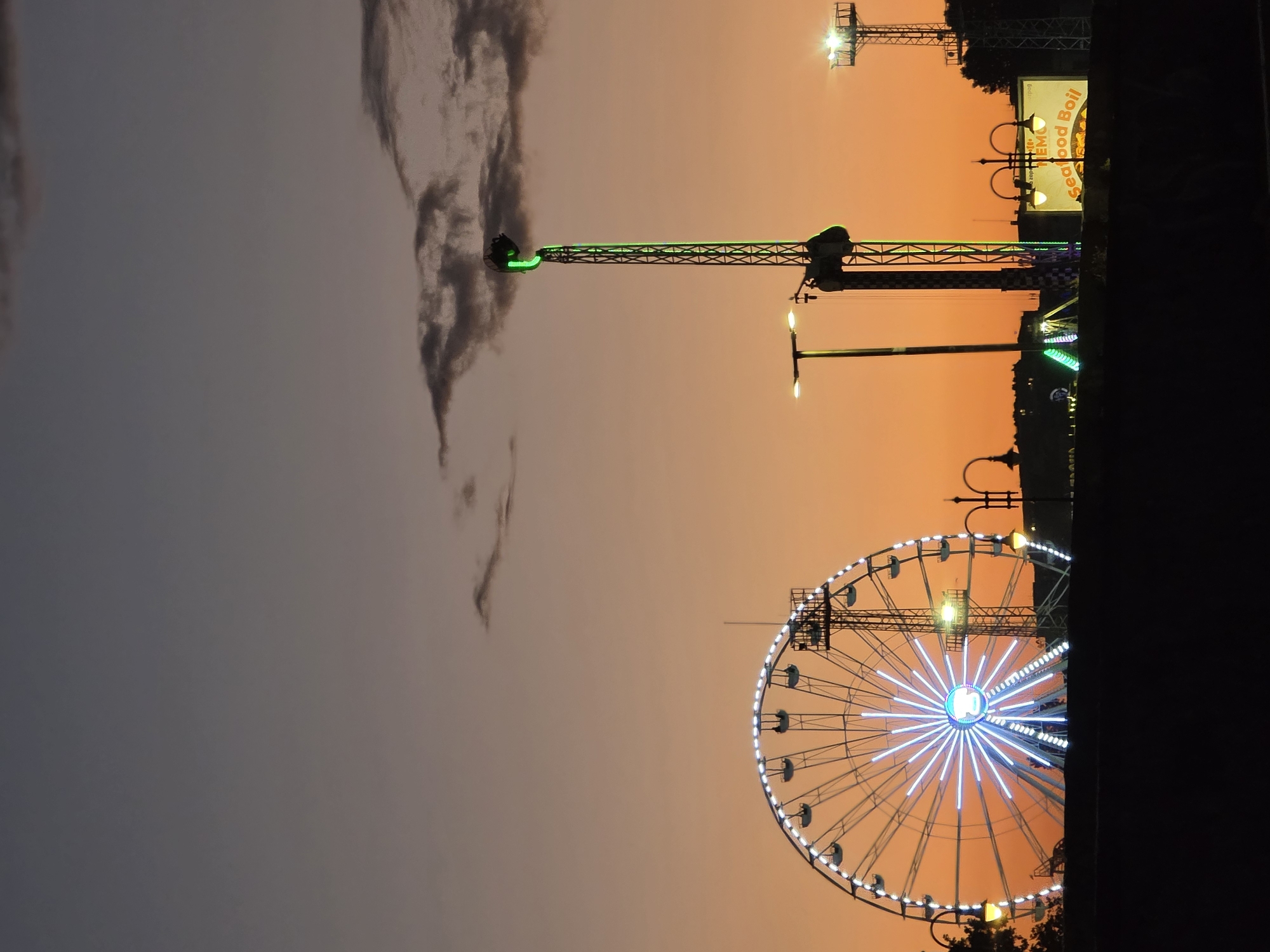 An amusement park is depicted at dusk with a Ferris wheel on the left and a drop tower on the right. The sky is a gradient of dark gray at the top fading to orange at the bottom. Clouds are visible in the upper portion of the sky. The Ferris wheel is illuminated with white lights and has multiple gondolas. The drop tower features a green illuminated top section and a dark structure extending upwards. Other amusement park rides and lights, including street lamps, are visible in the background. A billboard with "Seafood Boil" written on it is also present. The scene appears to be the Grand Mall amusement park in Varna, Bulgaria.