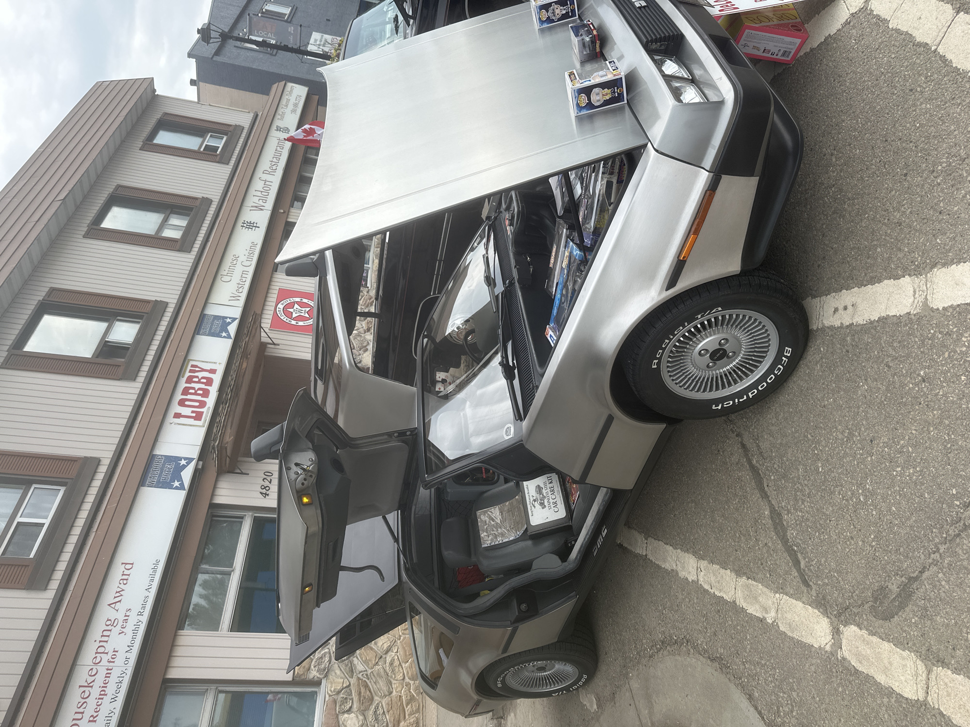 A silver DeLorean DMC-12 sports car with its gull-wing doors and hood open is parked on an asphalt street in front of a two-story building with a brick lower facade and wood-paneled upper facade. The building has signs indicating "Housekeeping Award," "WALDORF HOTEL," "LOBBY," and "Chinese Western Cuisine Waldorf Restaurant." The DeLorean's interior is visible, showing gray leather seats and a "Keep Your DeLorean Stainless Steel CAR CARE KIT" box. The engine compartment is open, revealing various items and products. Several Funko Pop figures and other merchandise are displayed on the edge of the opened hood. A Canadian flag is attached to the building's exterior above the restaurant sign. The street is marked with white lines, and there are some cracks and stains on the asphalt. Other vehicles and a lamppost are visible in the background. The scene appears to be an outdoor event or car show in the City of Leduc, Alberta, Canada, based on the context of the "WALDORF HOTEL" and nearby businesses.