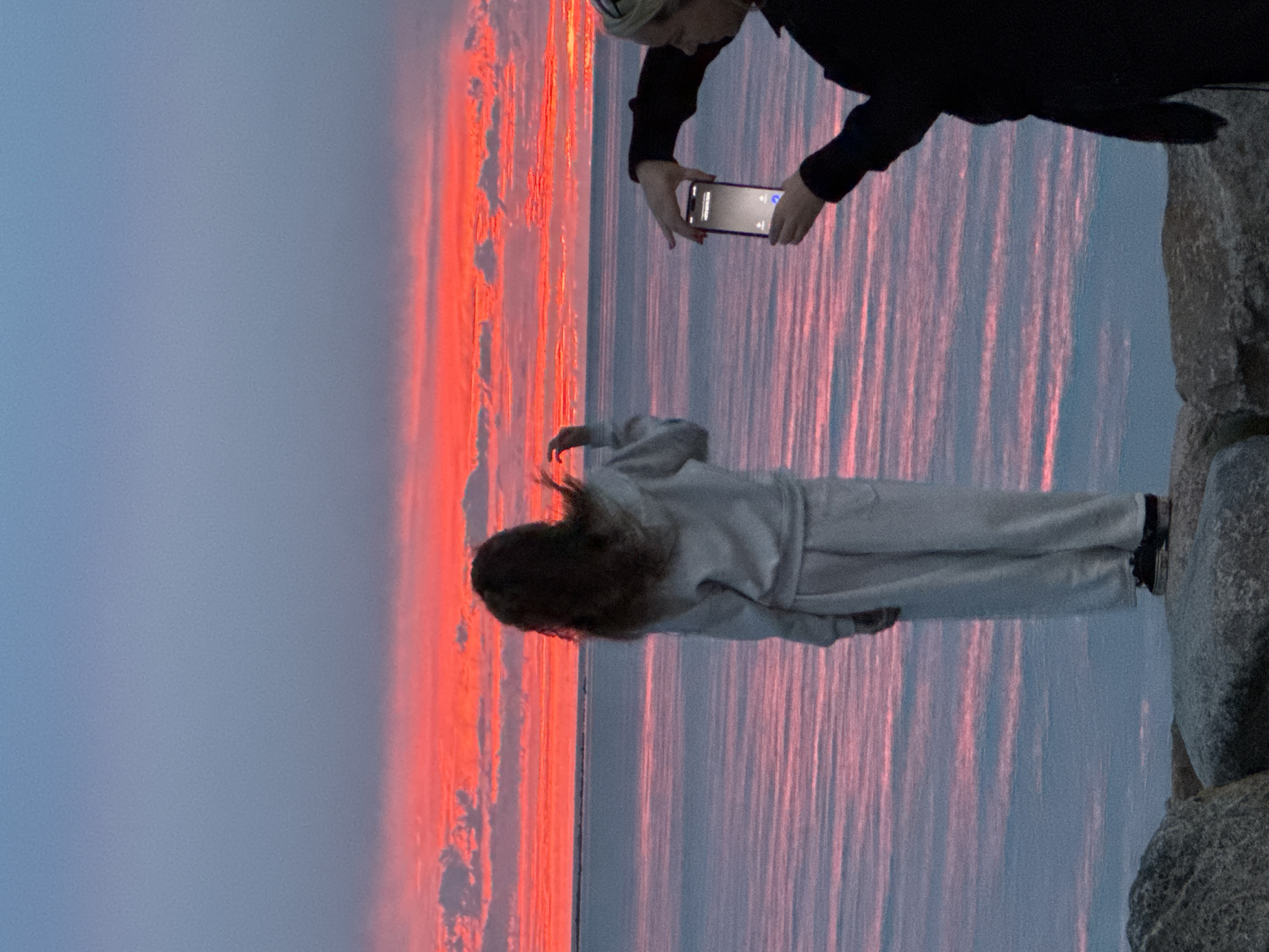 Two people stand by the water's edge, silhouetted against a vibrant sunset. The sky is a dramatic canvas of deep red and pink, with streaks of pale blue clouds. The water reflects the fiery sky, creating a shimmering, colorful expanse. In the foreground, one person, facing away, watches the sunset, while another to their right holds up a phone, likely capturing the breathtaking scene. Rocks line the shore, adding a natural texture to the composition.