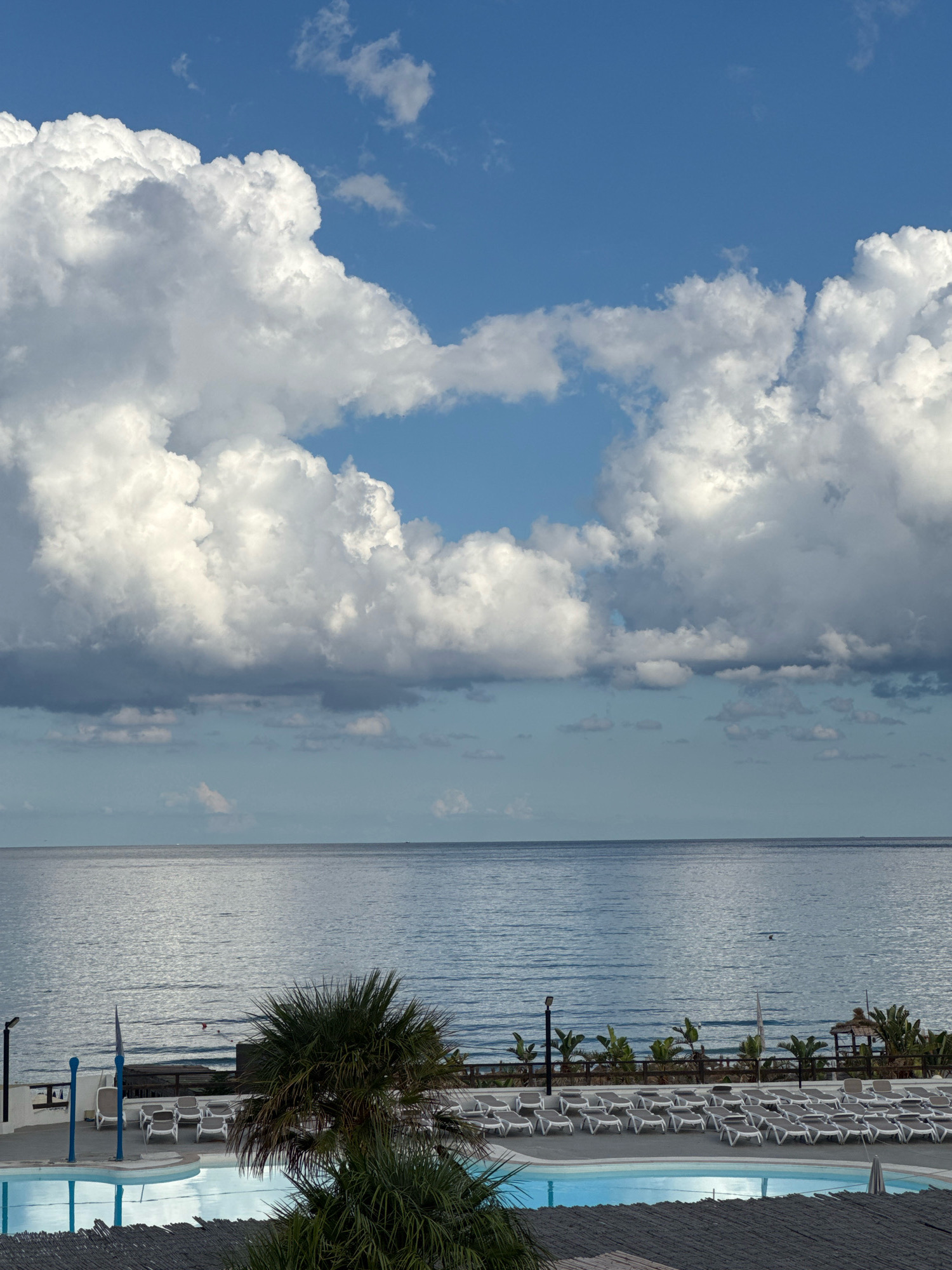 A serene scene unfolds with a vast expanse of calm blue ocean meeting a bright sky filled with dramatic white cumulus clouds. In the foreground, a resort setting features a sparkling blue swimming pool, surrounded by rows of empty white lounge chairs. Palm trees and other lush greenery dot the landscape, hinting at a tropical or Mediterranean climate. The water reflects the soft light of the sky, creating a tranquil and inviting atmosphere. There are no people visible in the image, giving the impression of a peaceful, deserted getaway. This image likely depicts a hotel or resort in Capaci, Italy, known for its beautiful coastal scenery and vacation spots.