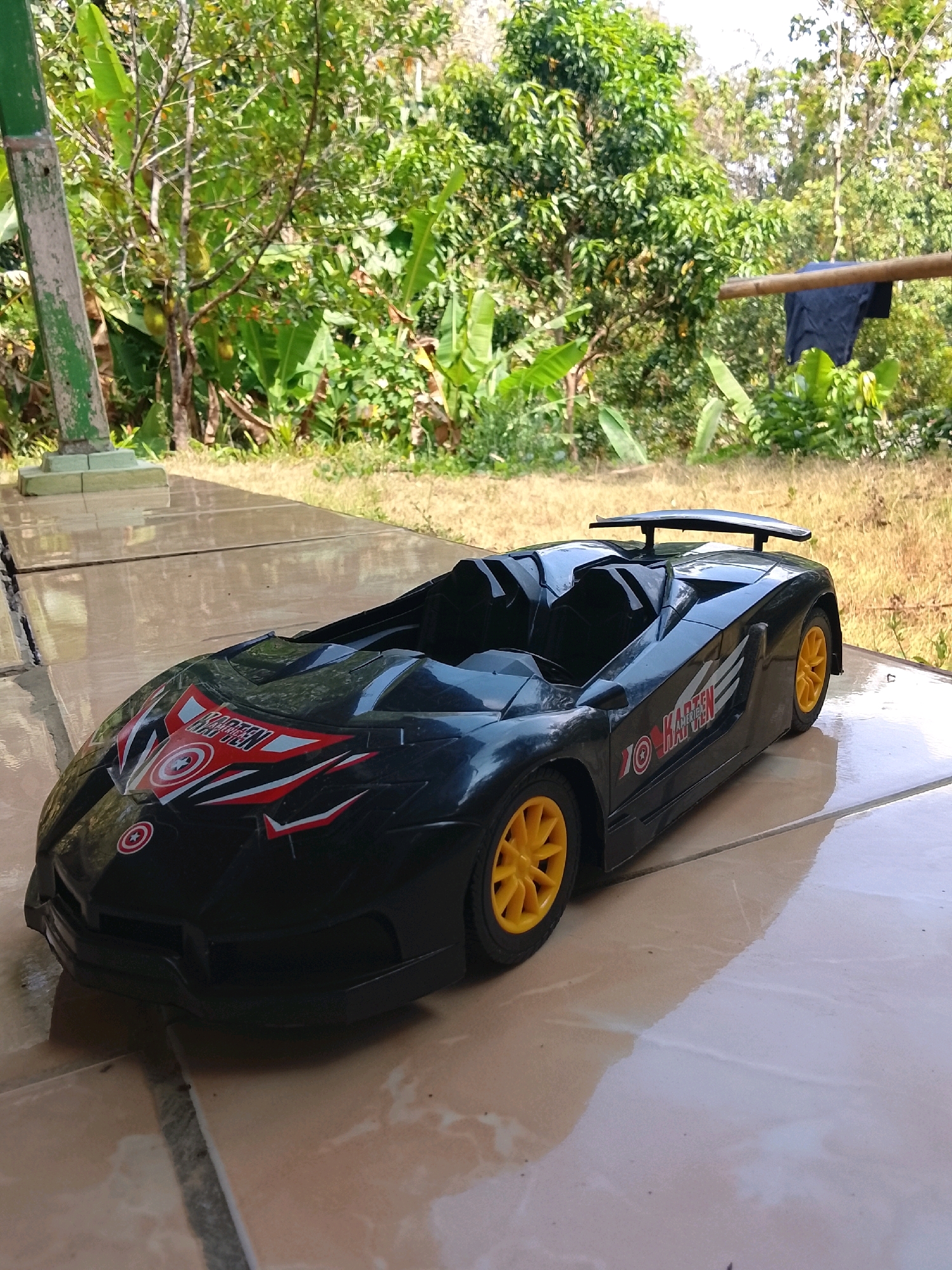 In Kertahayu, Indonesia, a black toy convertible car with yellow wheels is positioned on a tiled surface outdoors. The car features decorative decals, including a red and white design with a Captain America shield emblem on the hood, and text that appears to say "KARTEN MAJIKAN" on the side. The background consists of lush green vegetation, including trees and banana leaves, under a bright sky. To the right, a simple clothesline made of bamboo holds a dark blue garment. A green pillar, possibly part of a structure, is visible on the left side of the image. The tiled ground is wet, reflecting the surrounding environment.