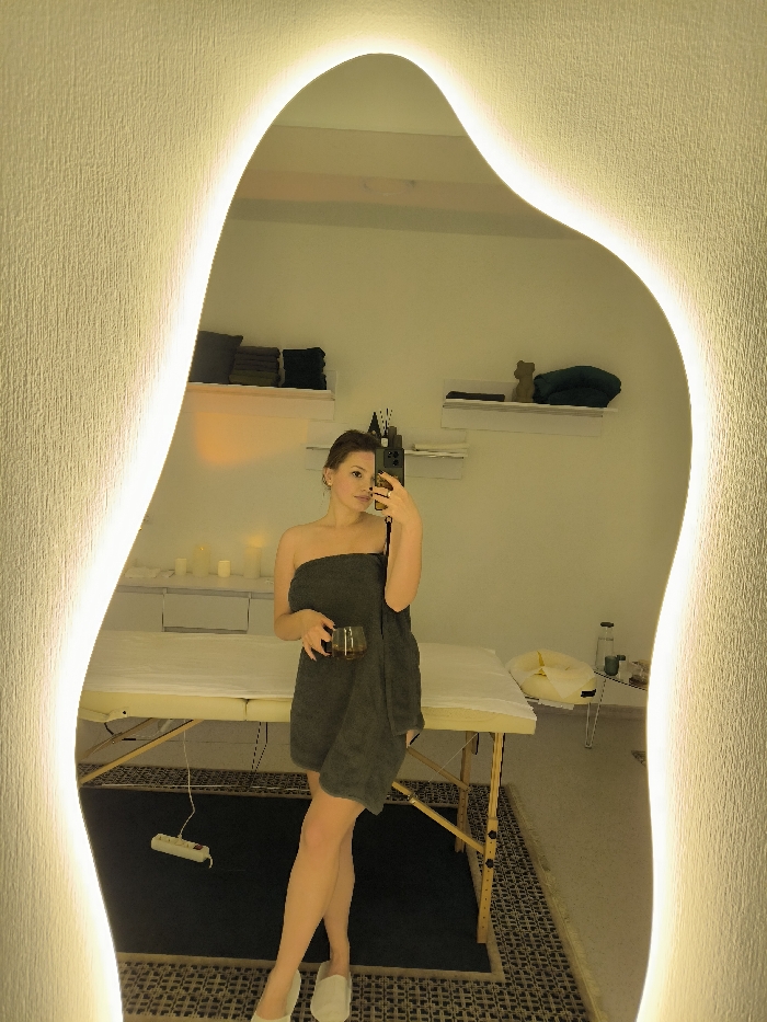 A young adult female is captured in a full-length mirror selfie within a spa or massage therapy room in Vinnytsia, Ukraine. She stands facing the mirror, holding a dark-colored glass in her left hand and a smartphone in her raised right hand, indicating she is taking the photograph. She is wrapped in a dark grey strapless towel, reaching above her knees, and wears light-colored slippers. The mirror is irregularly shaped with integrated warm backlighting, reflecting a room with light-colored, textured walls and ambient illumination.
Behind the female, a beige-framed massage table is draped with white linens. To the left, a white cabinet displays several white candles. Above this cabinet, a wooden shelf holds stacked dark green and grey towels. To the right, another white shelf holds a small decorative item resembling a bear and a folded teal towel. Further to the right, a small, white metal-framed side table supports a glass bottle, a small cup, and a light-colored, possibly yellow, basin or cushion. The floor is covered by a dark patterned rug, on which a white power strip with multiple outlets is visible near the base of the massage table.