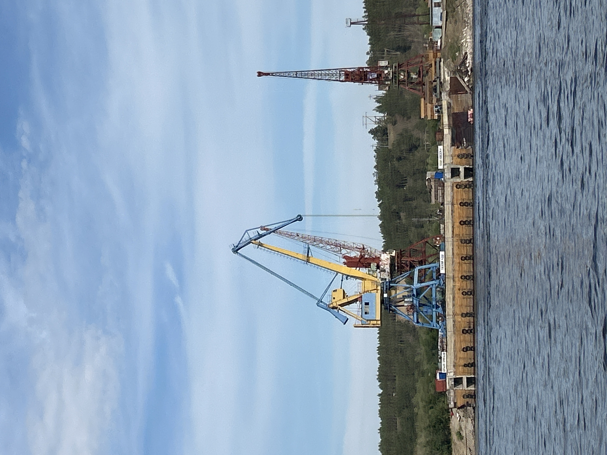The image depicts a waterfront scene in Nizhny Bestyakh, Russia, featuring industrial equipment along the shore of a wide river. In the foreground, dark blue water with visible ripples occupies the lower portion of the frame. Dominating the mid-ground is a large, tan-colored concrete structure, likely a dock or pier, with multiple cylindrical fenders attached along its edge. Several shipping containers and other industrial materials are visible on top of this structure.
Two prominent cranes stand on the dock. The larger crane on the left is a multi-colored gantry crane with a blue base, a yellow boom, and a red counterweight section. Its long boom extends upwards and to the right. To the right of this crane is a taller, slender red lattice boom crane, positioned further back on the shore. Its boom is oriented vertically.
Behind the dock and cranes, a dense line of dark green trees forms a backdrop, suggesting a forested area. The sky above is a bright blue with scattered white clouds. In the distance, on the right side of the image, a small structure with a tall pole or antenna is visible. The overall impression is of an active industrial port or shipyard. No people are discernible in the image.