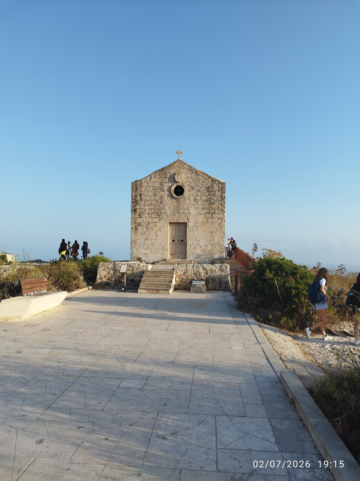 This media file captures a serene scene in Dingli, Malta. The focal point is a rustic stone chapel with a simple wooden door and a cross atop its gable. The chapel is situated on a rocky outcrop, overlooking a hazy expanse, likely the sea. The foreground features a paved walkway with an angular geometric pattern, and a white stone bench. To the left of the chapel, a group of silhouetted figures stand near a low stone wall, seemingly observing the view. To the right, two young women, one with a backpack, walk along a path bordered by dry vegetation. A wooden ramp leads up to the chapel from this side. The sky is a clear, bright blue, suggesting a pleasant day. The lighting indicates it is late afternoon or early evening, as evidenced by the long shadows cast across the paving stones and the warm glow on the chapel. The overall mood is peaceful and contemplative, evoking a sense of timelessness and natural beauty. Visible text in the lower right corner reads "02/07/2026 19:15", indicating the date and time the image was captured.