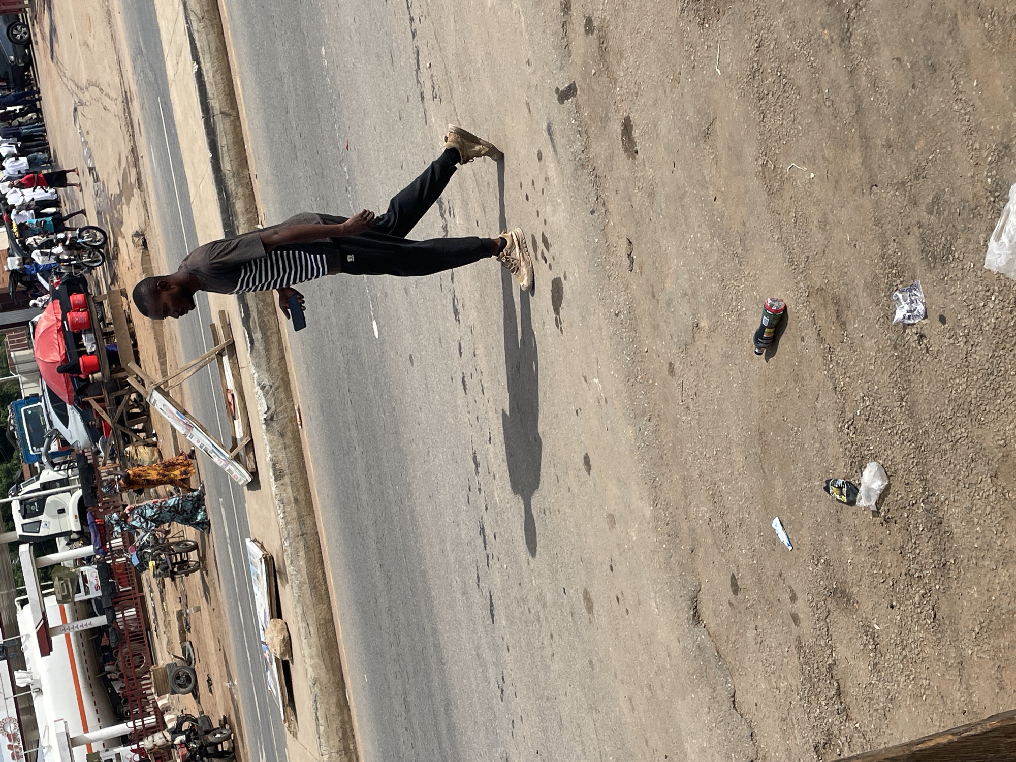The image shows a person walking along a paved road in Akwanga, Nigeria. The individual, wearing a striped shirt, dark pants, and light-colored sneakers, appears to be holding a mobile phone. The road is lined with parked vehicles, including trucks and motorcycles, alongside a variety of commercial establishments. Litter, including a bottle and wrappers, is scattered on the ground near the road's edge.