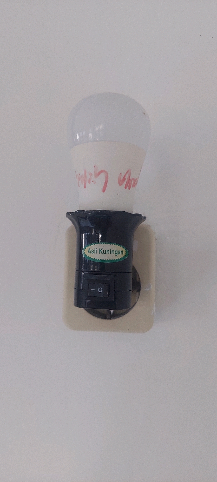 An LED light bulb is screwed into a black socket adapter with a built-in switch. The adapter is plugged into a standard electrical outlet on a white wall. The light bulb has red writing on it, but the text is illegible. A green oval sticker on the adapter reads "Asli Kuningan". The scene appears to be indoors, likely in a residential or commercial setting in Lumajang, Indonesia. The lighting is even, suggesting ambient indoor lighting.