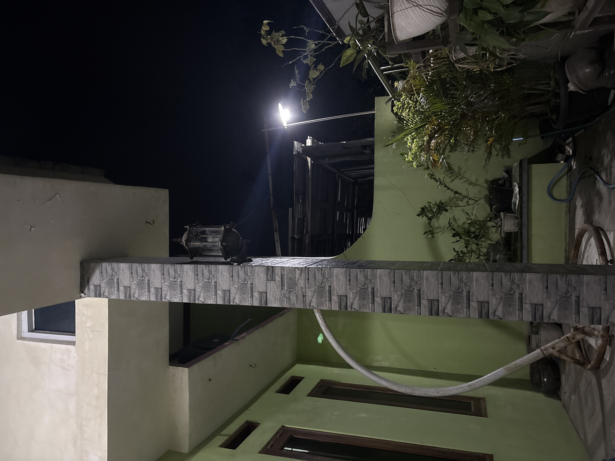 This photograph captures an outdoor scene at night, likely in the city of Gempol, Indonesia, showcasing a residential or commercial building's exterior. A light-colored, possibly stucco, upper story overhangs a pale yellow, textured wall on the left. Below this, a green wall with two dark-framed windows and two horizontal vents is visible. A white, curved pipe, possibly a drainpipe or conduit, extends from the green wall and curves down towards the ground.
A prominent rectangular pillar, clad in grey stacked stone veneer, stands in the center of the frame, dividing the scene. Attached to the pillar, or an adjacent structure, is an old-fashioned, dark-colored lantern with decorative metalwork, suspended from above.
To the right of the pillar, the green wall continues, featuring various potted plants. Among these are a larger palm-like plant in a dark pot and other foliage, some of which appear to be illuminated by a nearby street light. The street light itself is a modern, white fixture mounted on a pole, casting a bright, diffused light into the dark night sky. In the background, parts of a sloped roof structure are visible, possibly an extension or an adjacent building. The ground appears to be paved, with some hoses visible near the base of the pillar and plants. The overall ambiance is nocturnal and functional, highlighting architectural elements and the surrounding vegetation under artificial light.