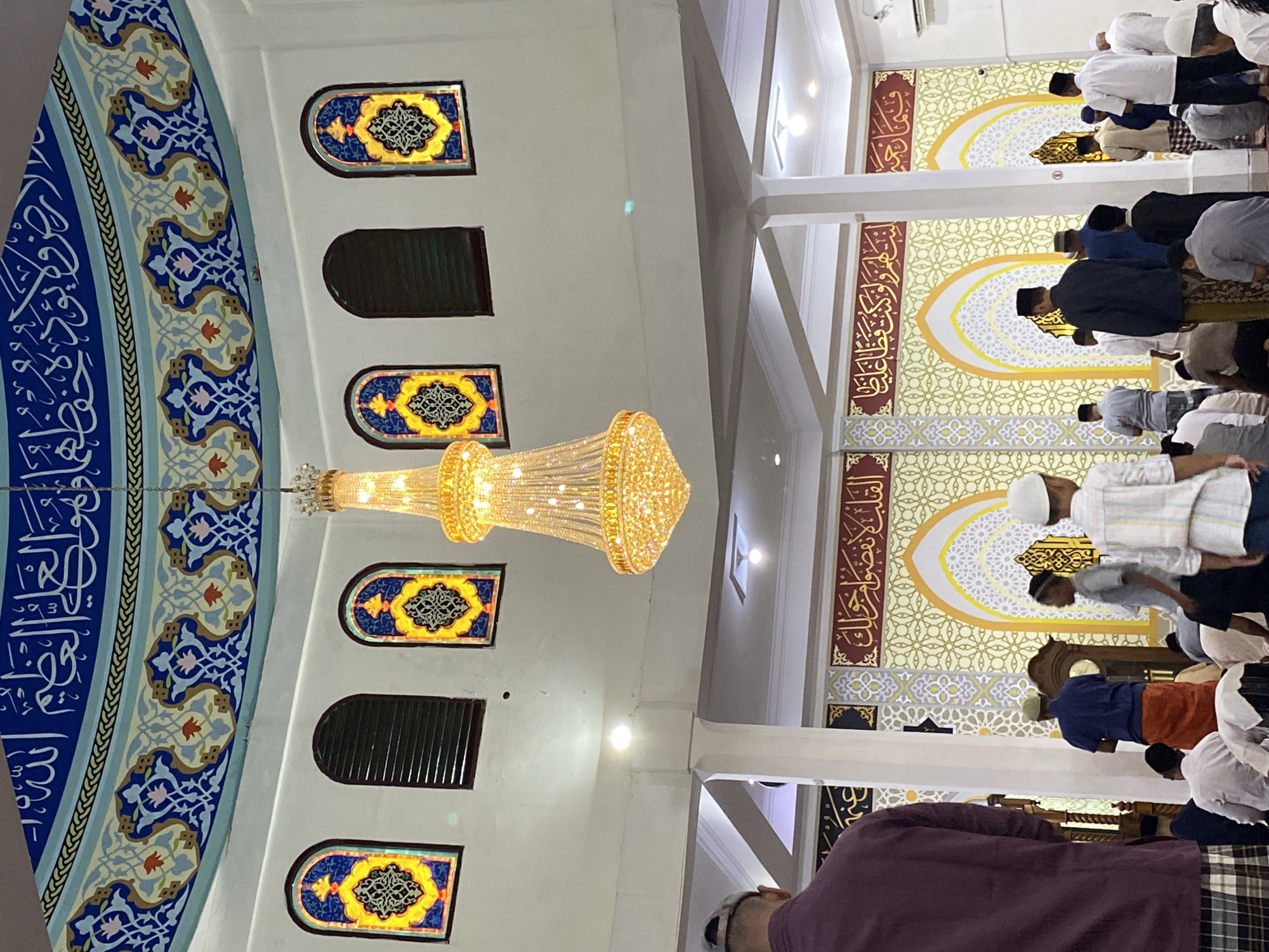 The image depicts the interior of the Islamic Center mosque in Lhokseumawe, Indonesia. A large, ornate chandelier hangs from the domed ceiling, which is decorated with intricate blue and gold Islamic calligraphy and patterns. Arched windows with stained-glass inserts featuring geometric designs are visible along the upper walls. Below, a group of men and boys, some wearing white skullcaps, are gathered for prayer. The walls are adorned with decorative panels featuring more Islamic calligraphy and geometric motifs in yellow, red, and white. A man in a maroon shirt and plaid pants stands in the foreground, facing away from the camera. Another man in a blue shirt and orange sarong is visible in the mid-ground, facing the wall. The overall atmosphere suggests a place of worship during a prayer service.