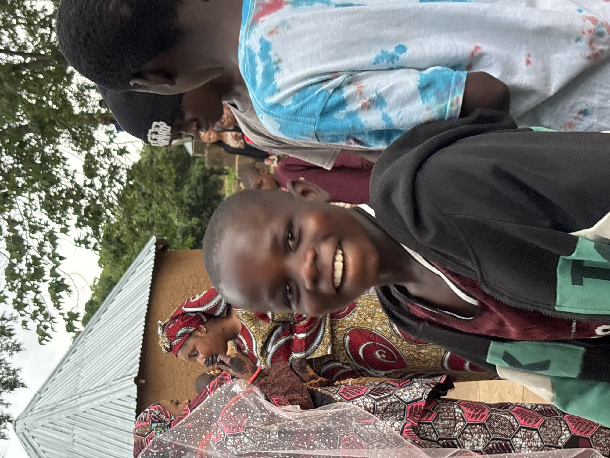 A group of people are gathered outdoors. In the foreground, a young boy with a wide smile looks directly at the camera. He is wearing a dark hooded jacket over a maroon shirt. To his left, a woman in a colorful patterned dress and headscarf is speaking to someone. Behind them, other individuals are visible, some with their backs to the camera. The background features a building with a corrugated metal roof and trees. The scene appears to be in Jos, Nigeria, indicated by the style of dress, architecture, and general environment.