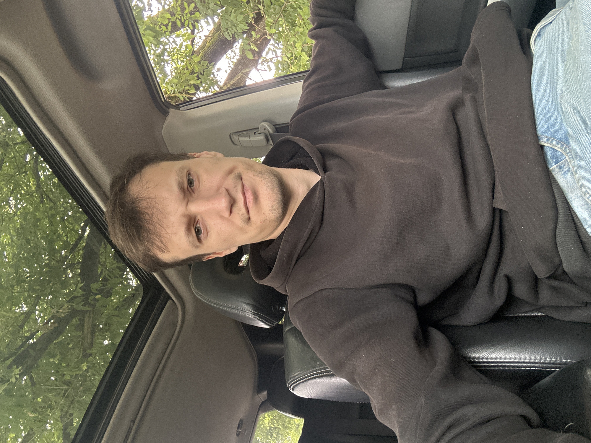 A young man with short brown hair and a slight stubble is sitting in the driver's seat of a vehicle. He is wearing a dark brown hooded sweatshirt and blue jeans. The man is looking directly at the camera with a neutral expression, with his head tilted slightly to his left. His left arm is resting on the center console and his right arm is resting on the car door. The vehicle has a sunroof, which is open, revealing green trees and foliage above. The interior of the car is dark, with black leather seats visible. The scene appears to be outdoors, likely in a wooded area, possibly on the outskirts of Makiv, Ukraine, given the presence of trees and the general atmosphere.