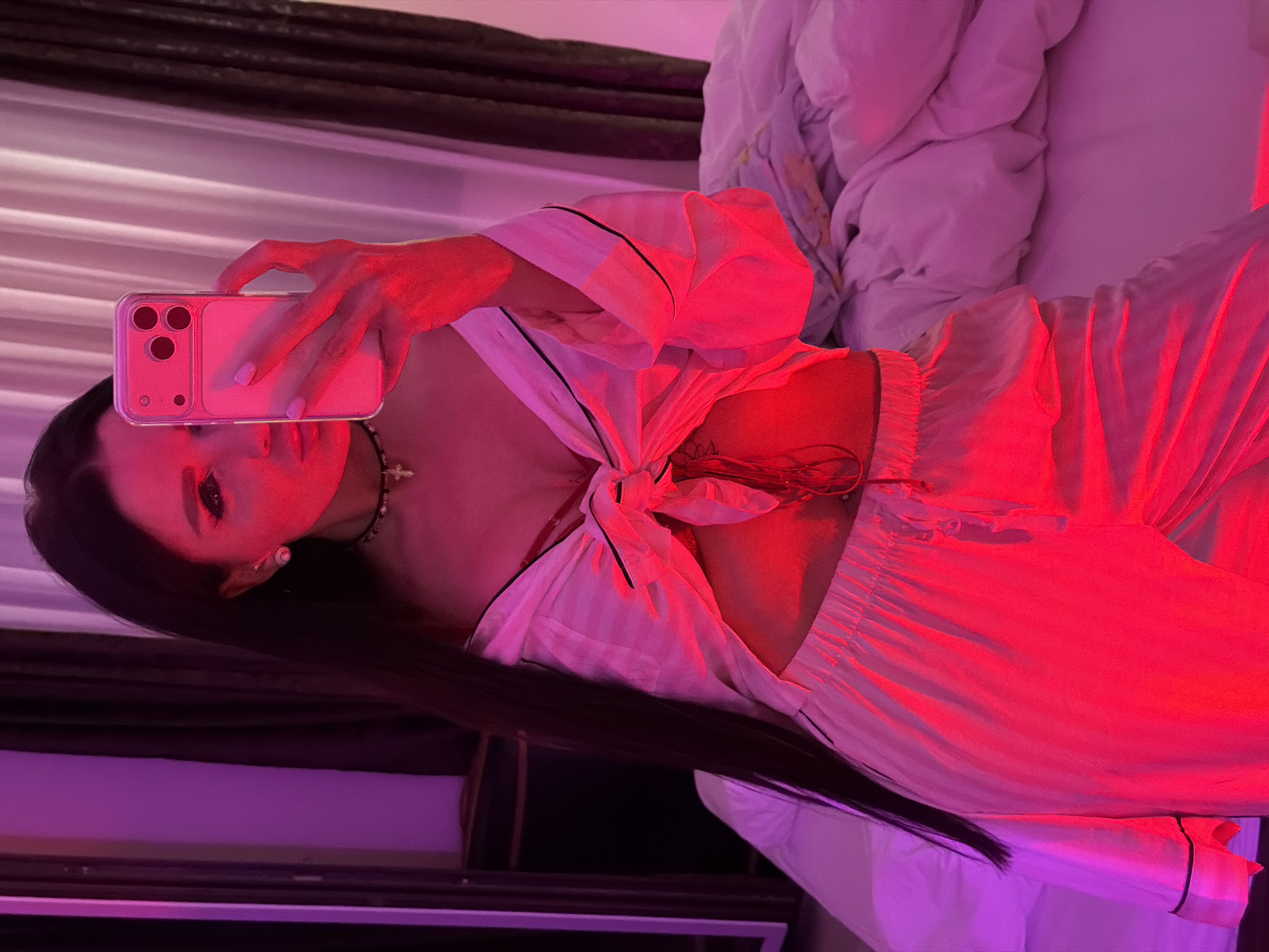 A person is captured in an indoor mirror selfie, likely lying down, with the scene illuminated by striking artificial red and purple lighting. The setting appears to be a bedroom or lounge area in Boreti, Montenegro, with soft, light-colored bedding visible in the background.
The subject, a woman with long, dark hair, is holding a light-colored smartphone (identifiable by its triple-lens camera array and clear case, suggesting an iPhone) to take the photo. Her face is partially obscured by the phone and bathed in a red glow, with her eyes clearly visible. She wears light-colored (possibly white or cream) pajamas or loungewear featuring black piping. The top is tied at the front, creating a knotted detail and revealing a portion of her midriff and décolletage. A necklace with a small cross pendant is visible around her neck.
The right side of the image is dominated by intense red light, highlighting the subject's clothing and the immediate surroundings. The left side transitions to a softer purple light, illuminating the background elements which include horizontal patterns that could be a headboard or wall, and ruffled bedding. The overall atmosphere is intimate and moody due to the specific lighting choices. No discernible text is visible in the image.