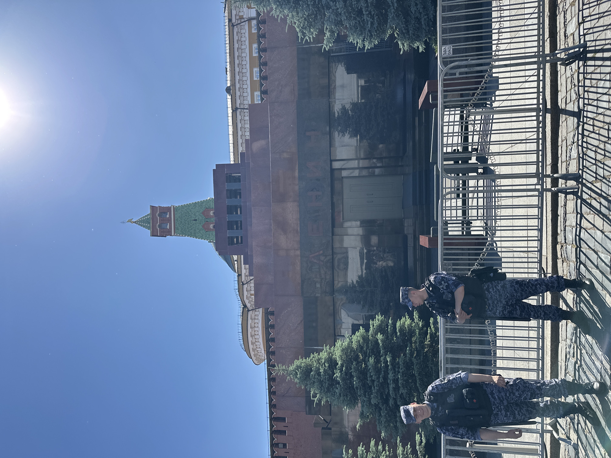 The image captures a scene in Red Square, Moscow, Russia, featuring the Lenin Mausoleum under a clear blue sky. The mausoleum, a tiered structure of dark red/brown stone, dominates the midground, with large Cyrillic letters spelling "ЛЕНИН" (LENIN) on its reflective facade. Behind the mausoleum, a segment of the red brick Kremlin wall and a green-roofed tower of the Kremlin with a golden spire are partially visible. Several coniferous trees are positioned in front of the mausoleum. In the foreground, two men in blue/grey camouflage uniforms, tactical vests, and matching caps stand behind silver metal crowd control barriers. The man on the left faces slightly towards the viewer, exhibiting a faint smile, while the man on the right faces away, his arms crossed over his chest. The ground is paved with cobblestones, and strong, defined shadows are cast across the foreground, indicating bright sunlight from a high angle.
