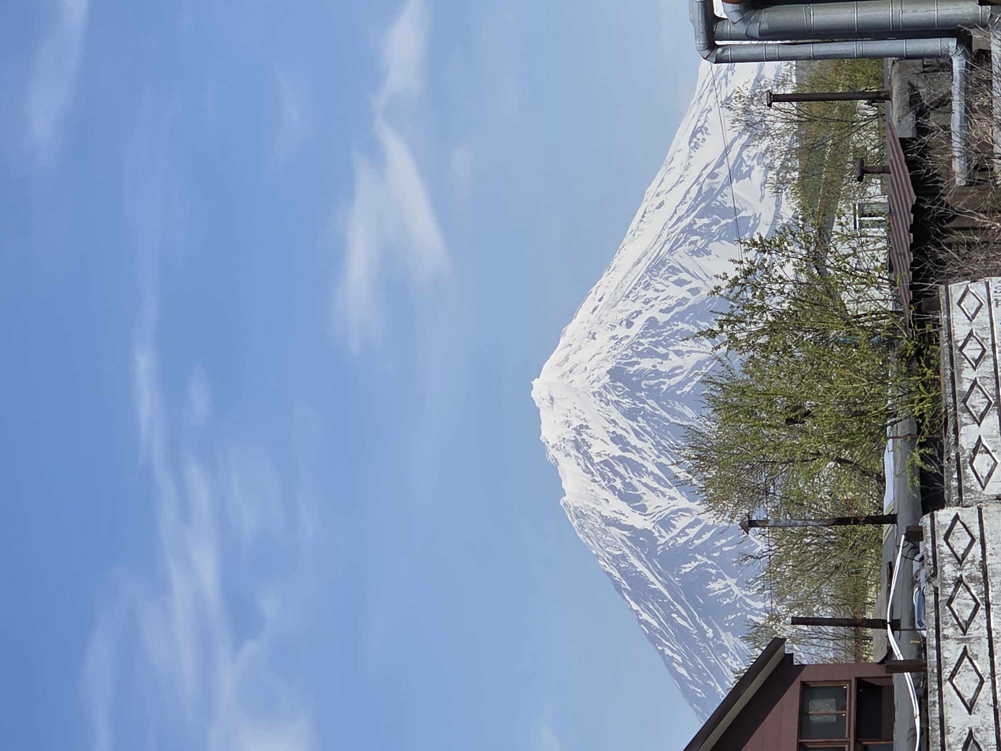 A vertical shot captures a majestic snow-capped mountain under a clear blue sky with sparse, wispy clouds. The mountain, a prominent stratovolcano, exhibits extensive snow cover on its summit and slopes, interspersed with darker rock or bare areas. In the foreground, from the left, a portion of a brown wooden house with multiple rectangular windows is visible. Below the house, a white concrete fence with a repeating pattern of dark, diamond-shaped depressions spans the width of the lower image. Above this fence, several dark horizontal pipes or cables are present. Numerous deciduous trees, some with emerging green leaves and others still bare, are scattered across the mid-foreground, partially obscuring the lower slopes of the mountain and other structures. Several dark vertical pipes or chimneys rise from behind the fence and trees. On the far right, a cluster of large, metallic, cylindrical pipes, possibly for utilities, ascends vertically, partially cropped by the image edge. The scene depicts a landscape view from a settled area in the city of Unknown, Unknown.