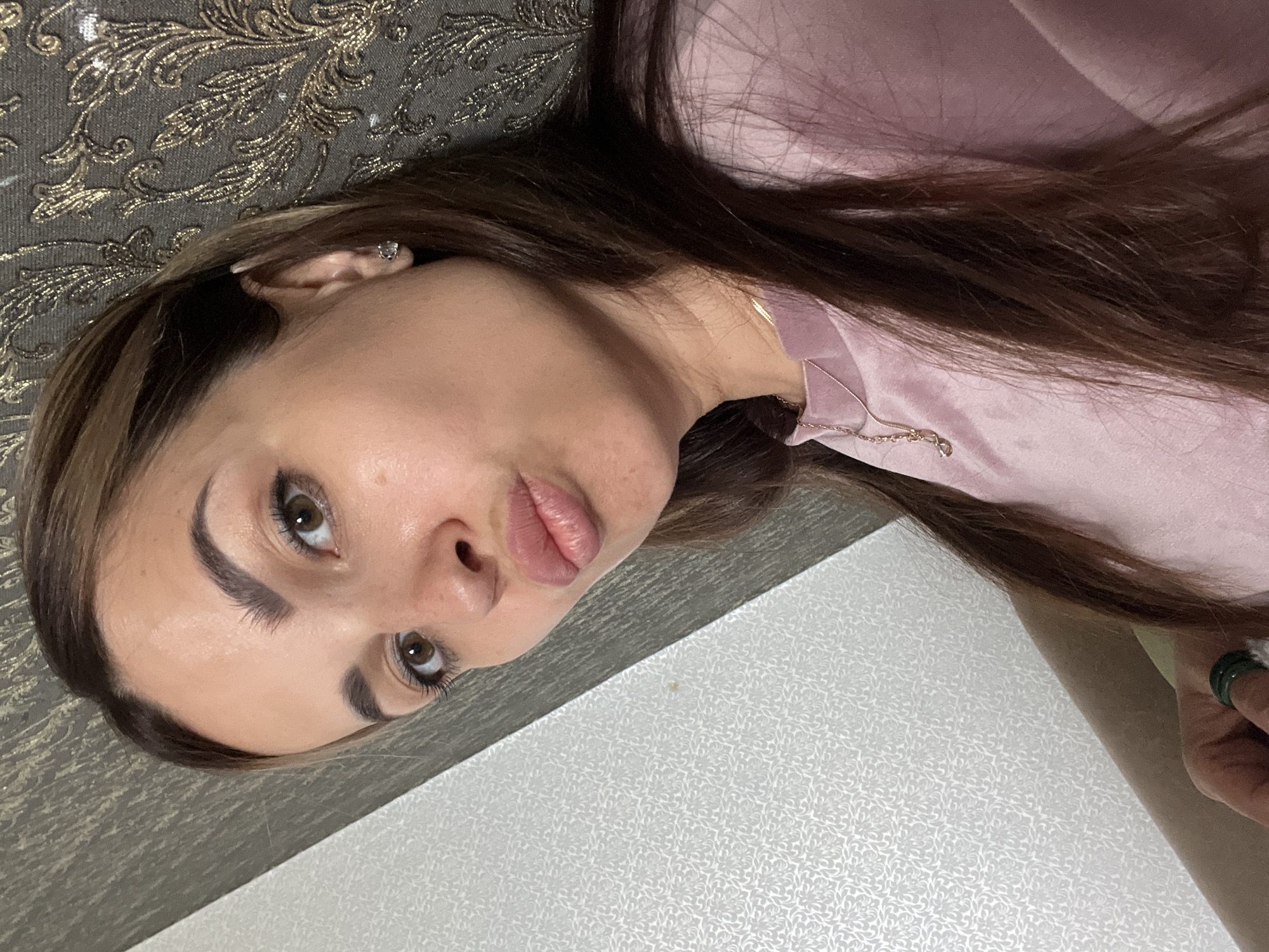 An overhead shot shows a woman with long, brown hair and brown eyes making a kiss face at the camera. She is wearing a pink shirt with a gold necklace and a green ring on her left hand. She is leaning against a wall with a gold patterned wallpaper. The wall to her left is white with a subtle pattern. The lighting appears to be artificial. The background suggests an indoor environment.