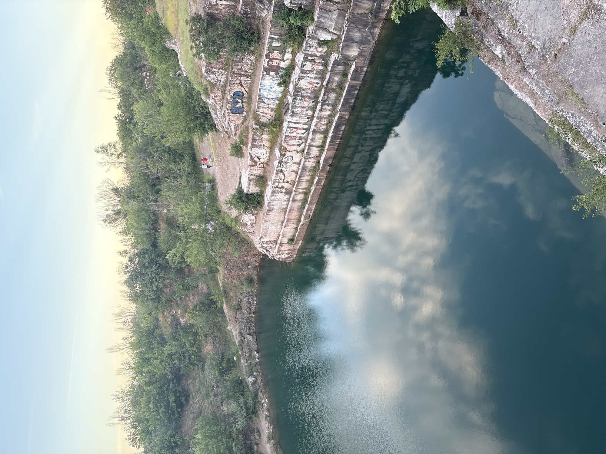 The image shows a body of water within a quarry. The quarry walls are terraced, with graffiti visible on some sections. Vegetation is present along the top edges of the quarry walls. The water reflects the sky. Two figures are visible on a ledge near the graffiti. The image's orientation is rotated 90 degrees clockwise. The location is likely Pravoslav, Bulgaria.