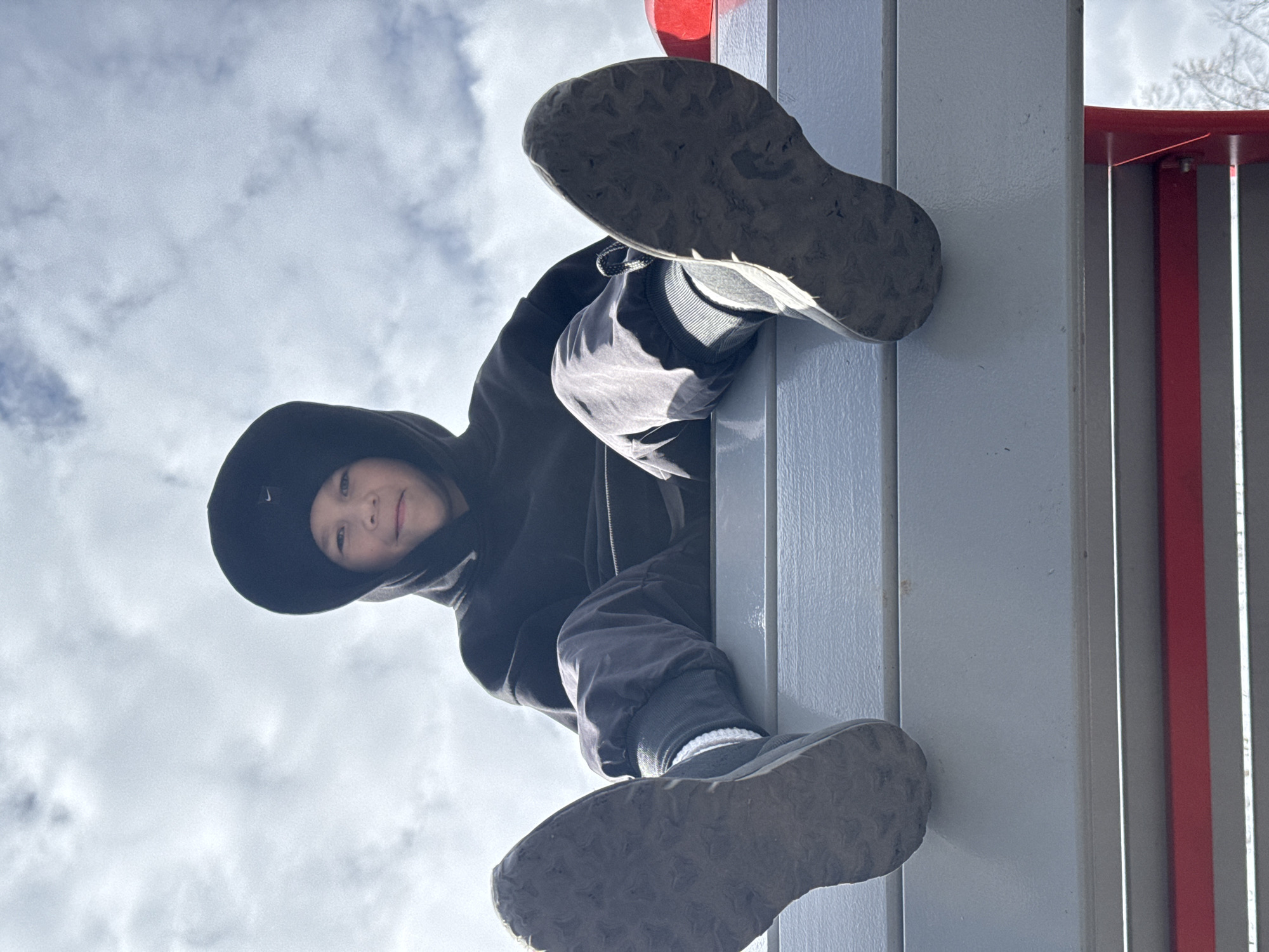A low-angle shot captures a child, possibly male, looking down at the camera from atop a structure. The child is dressed in a black hooded sweatshirt and dark pants. Their shoes, seen close-up in the foreground, have textured soles and light-colored socks peeking out. The structure appears to be part of a playground or outdoor climbing frame, with gray and red elements visible. The sky is visible in the background, showing a mix of clouds and blue sky. The photo does not contain sufficient information to establish a connection to Zav'yalovo, Russia, or any specific location.