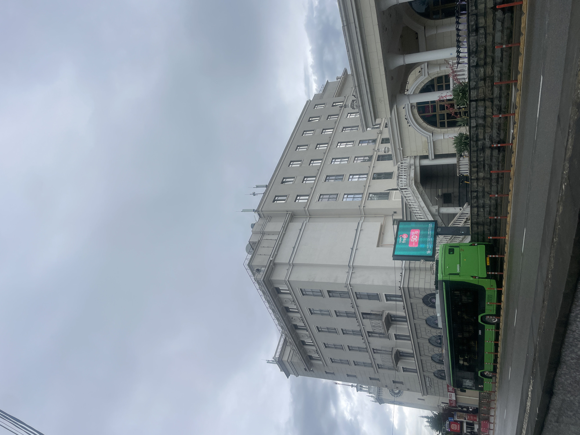 The image shows a large, light-colored building in what appears to be an urban setting. The building has multiple stories and a symmetrical facade with numerous windows arranged in a regular grid. Architectural details include decorative elements, balconies, and a roofline with distinct setbacks. Adjacent to the building is a green bus with a digital display screen, parked along the street. The screen shows an advertisement. The sky is overcast, suggesting either early morning, late afternoon, or a cloudy day. The location appears to be the city of Sochi, Russia. There are also some smaller building structures visible closer to the bus with signs that are advertising shops.