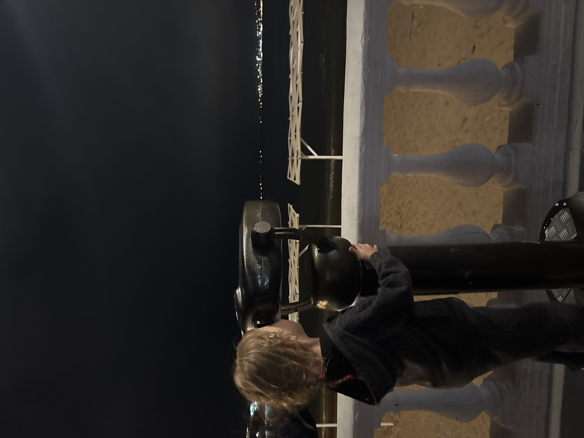At night, a young individual with blond hair looks through a coin-operated binocular telescope at a seaside location in what appears to be Gelendzhik, Russia. The person is positioned near a white stone balustrade, with a sandy area visible behind it. In the distance, along the dark body of water, a string of lights suggests a coastline. A white structure, possibly part of the beachfront infrastructure, stands beyond the balustrade. The scene is dimly lit, casting shadows and emphasizing the contrast between the dark night sky and the distant lights. The child is wearing a dark hooded sweatshirt.