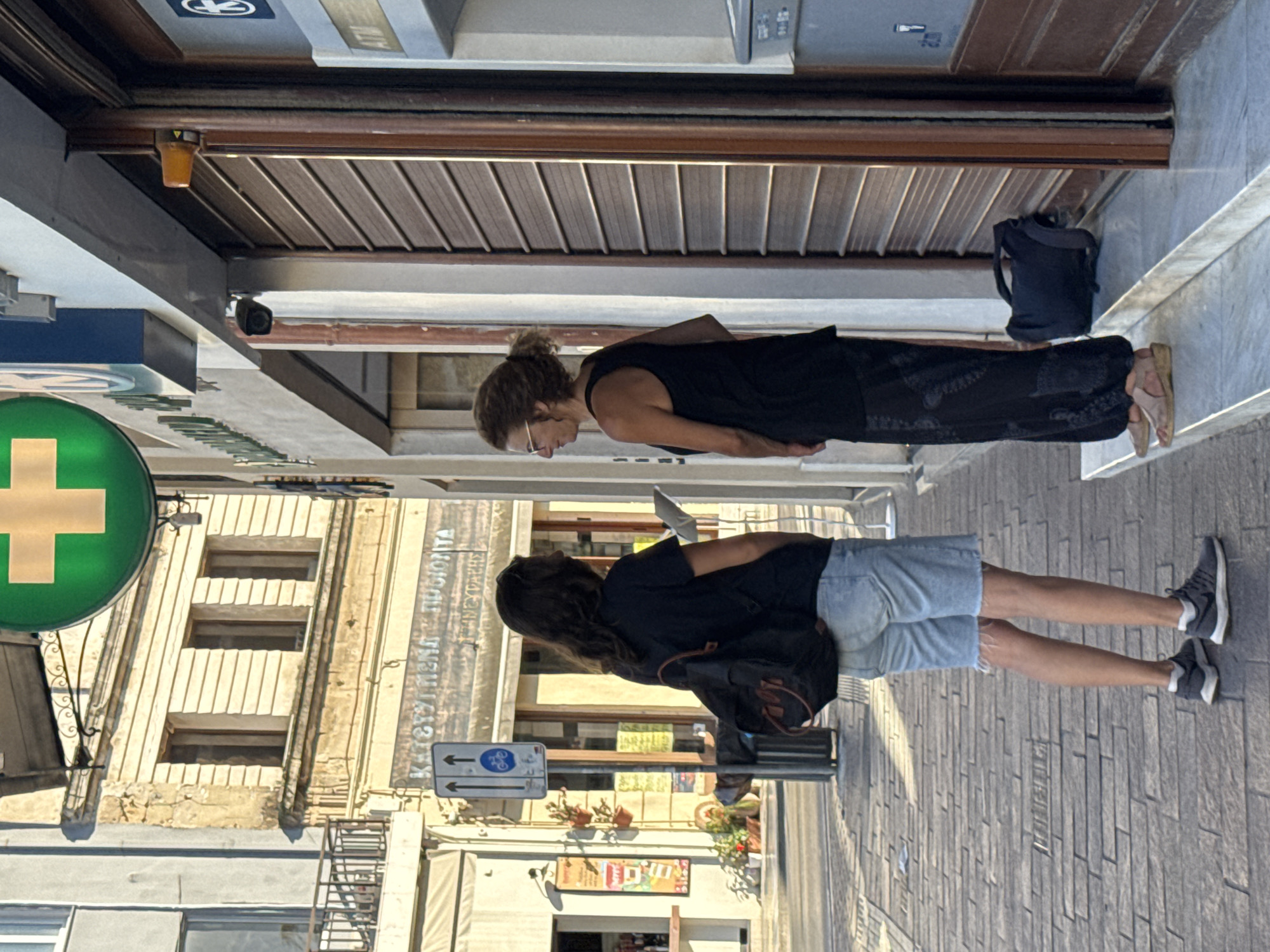 Two individuals are standing on a cobblestone sidewalk in front of a building on a street in Chania, Greece. The building appears to be a commercial establishment with a brown roll-up door and a green pharmacy sign with a white cross at the top. To the left of the entrance, there is an ATM machine.
The person on the left, facing away from the camera, has long, dark, wavy hair and is wearing a black t-shirt, light-wash denim shorts with frayed edges, and grey athletic shoes. They are carrying a black backpack with brown straps and appear to be holding something in their hands.
The person on the right, facing left and slightly towards the camera, has curly, light brown hair and is wearing glasses. They are dressed in a black tank top and loose-fitting black patterned pants. They are wearing tan sandals and have a dark bag placed on the steps in front of the building. Both individuals appear to be engaged in conversation or looking at something together.
The sidewalk is paved with grey cobblestones. In the background, there are buildings with architectural details characteristic of older European cities, including balconies and arched windows. A street sign indicating a pedestrian and bicycle path is visible near the center of the frame. The lighting suggests it is daytime, with shadows cast on the sidewalk.